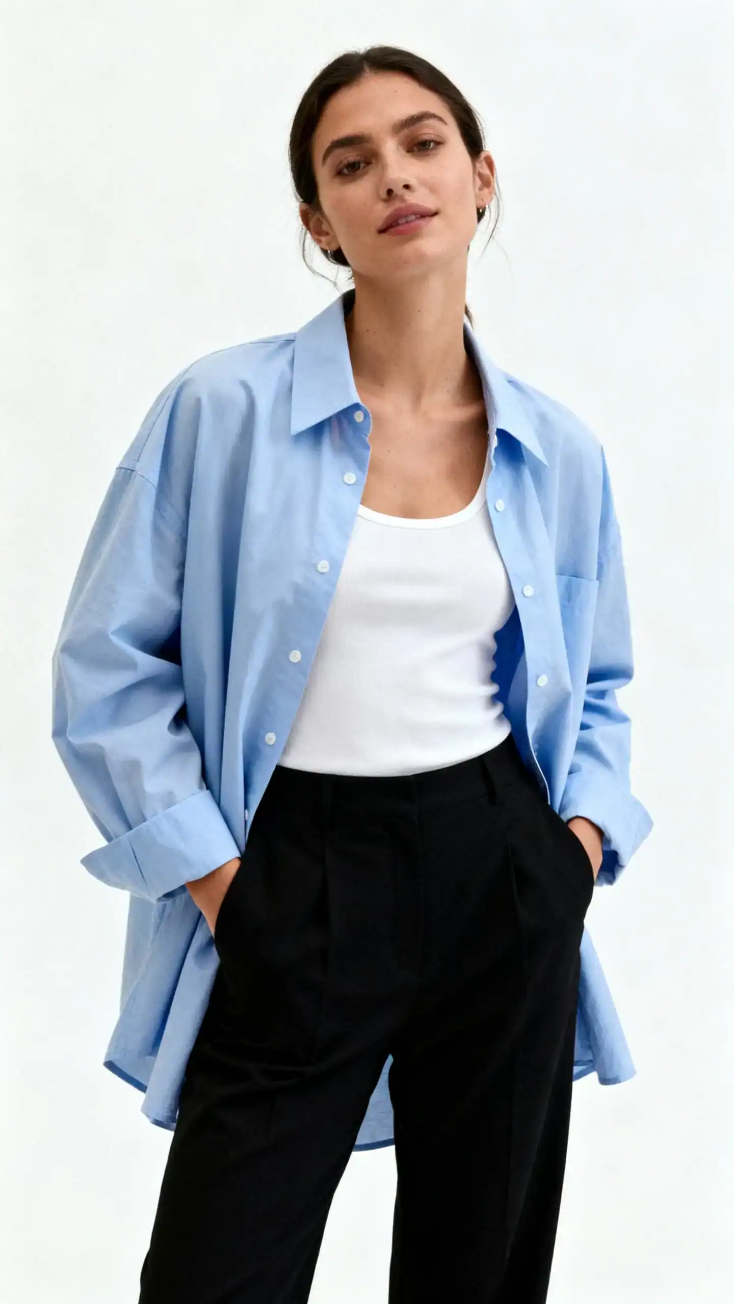 Editorial studio photo of a woman in black trousers with a minimalist white tank and an oversized light blue button-up shirt worn open, white background, studio lighting, relaxed hands-in-pockets pose, clean composition, cool and uncomplicated.