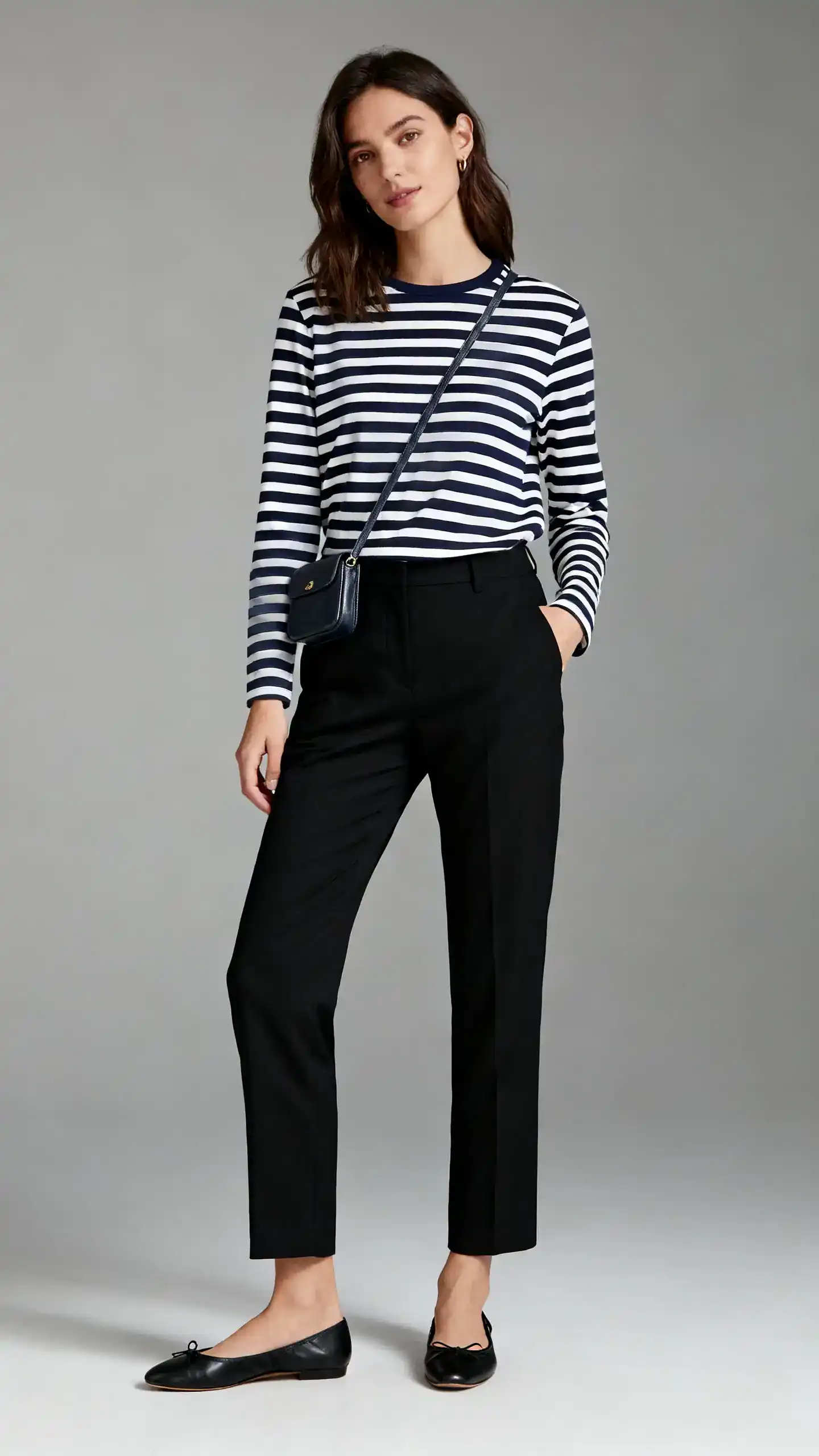 Editorial style photo of a woman in black trousers with a classic navy-and-white Breton stripe long-sleeve tee, simple black ballet flats and a petite crossbody, gray background, studio lighting, relaxed stance with one hand in pocket, clean composition, polished.