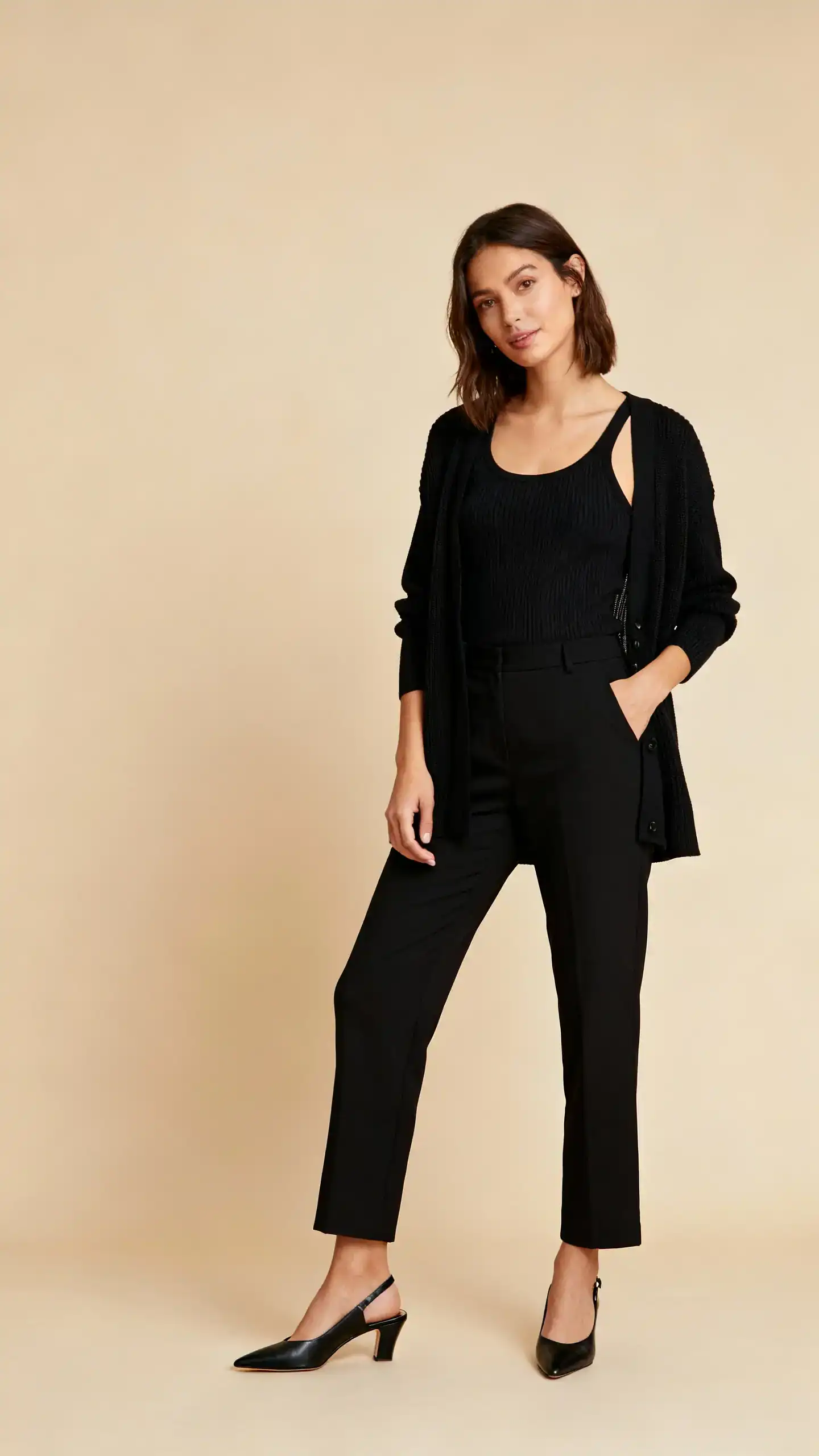 Editorial style photo of a woman in black trousers with a coordinated cardigan set (fine-gauge knit tank and matching cardigan) and sleek black slingback heels, beige background, studio lighting, relaxed standing pose, clean composition, casual-cool and polished.