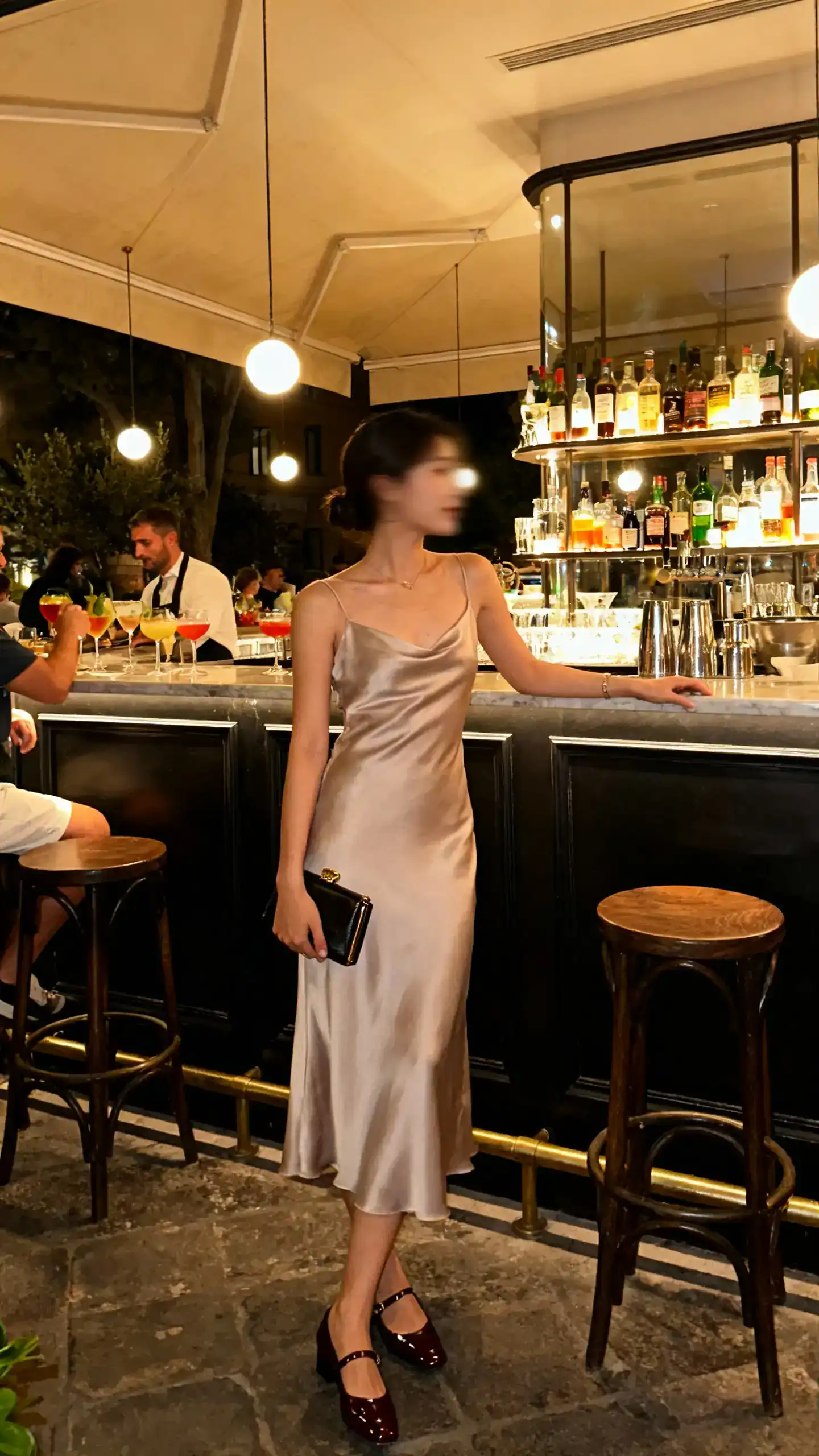 Evening candid of a woman wearing a slip dress with polished flats and a small clutch, standing at a bar during aperitivo, face slightly blurred, warm ambient light, iPhone photo quality.