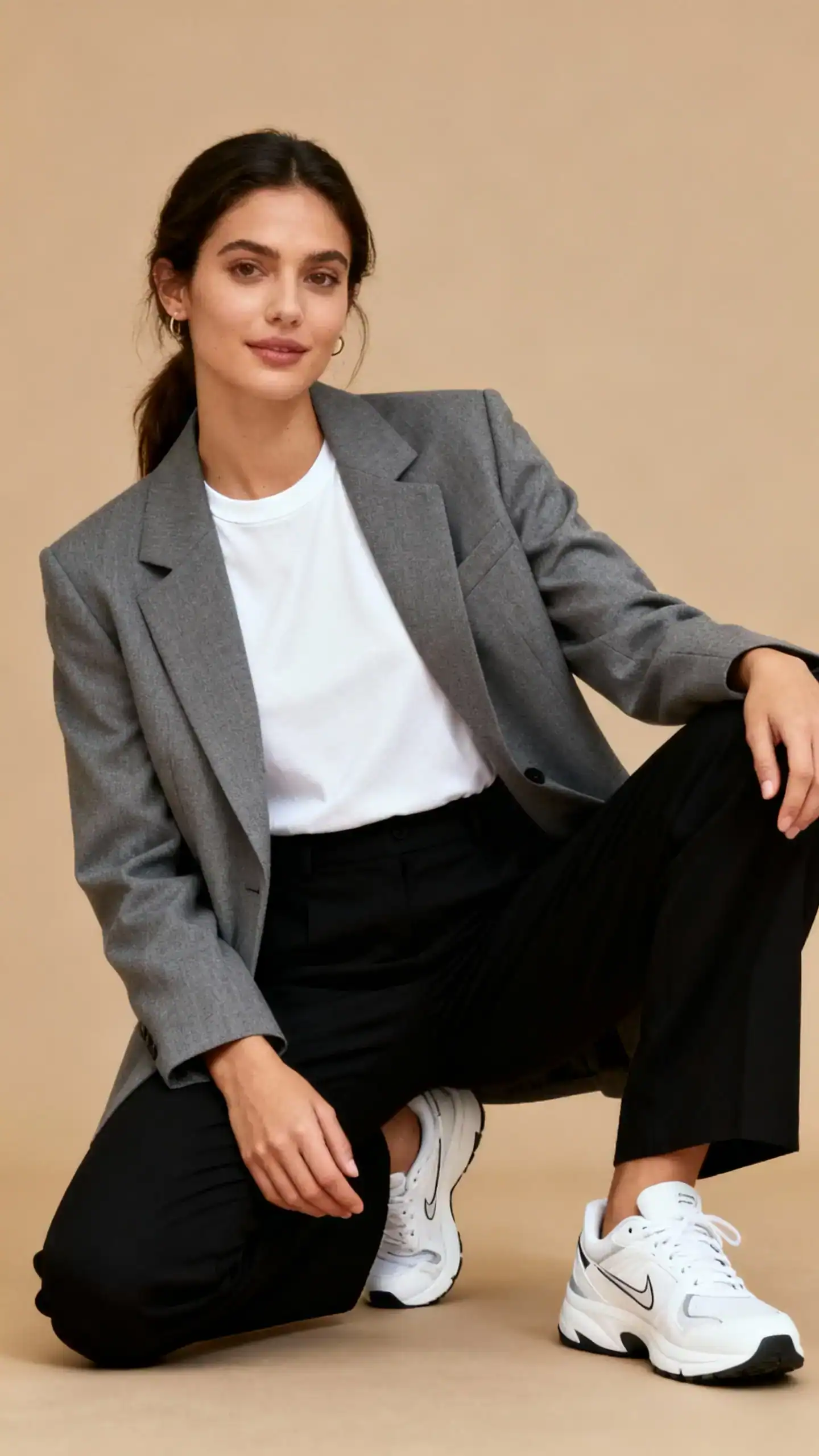 Studio fashion photo of a woman wearing black trousers with a structured gray blazer over a white crewneck tee and sporty white sneakers, tan background, soft studio lighting, casual-professional pose, clean composition, refined and modern.