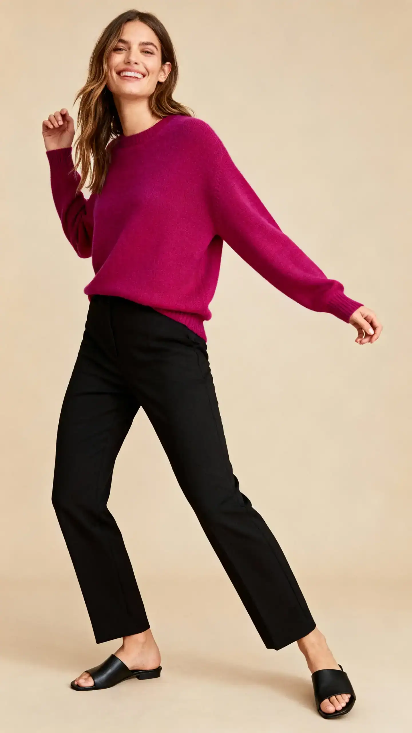 Studio fashion photo of a woman wearing black trousers with a bright pop-color knit (vibrant cobalt or cherry red crewneck) and minimalist black mules, sand-toned background, soft studio lighting, upbeat natural pose, clean composition, fresh and sleek.