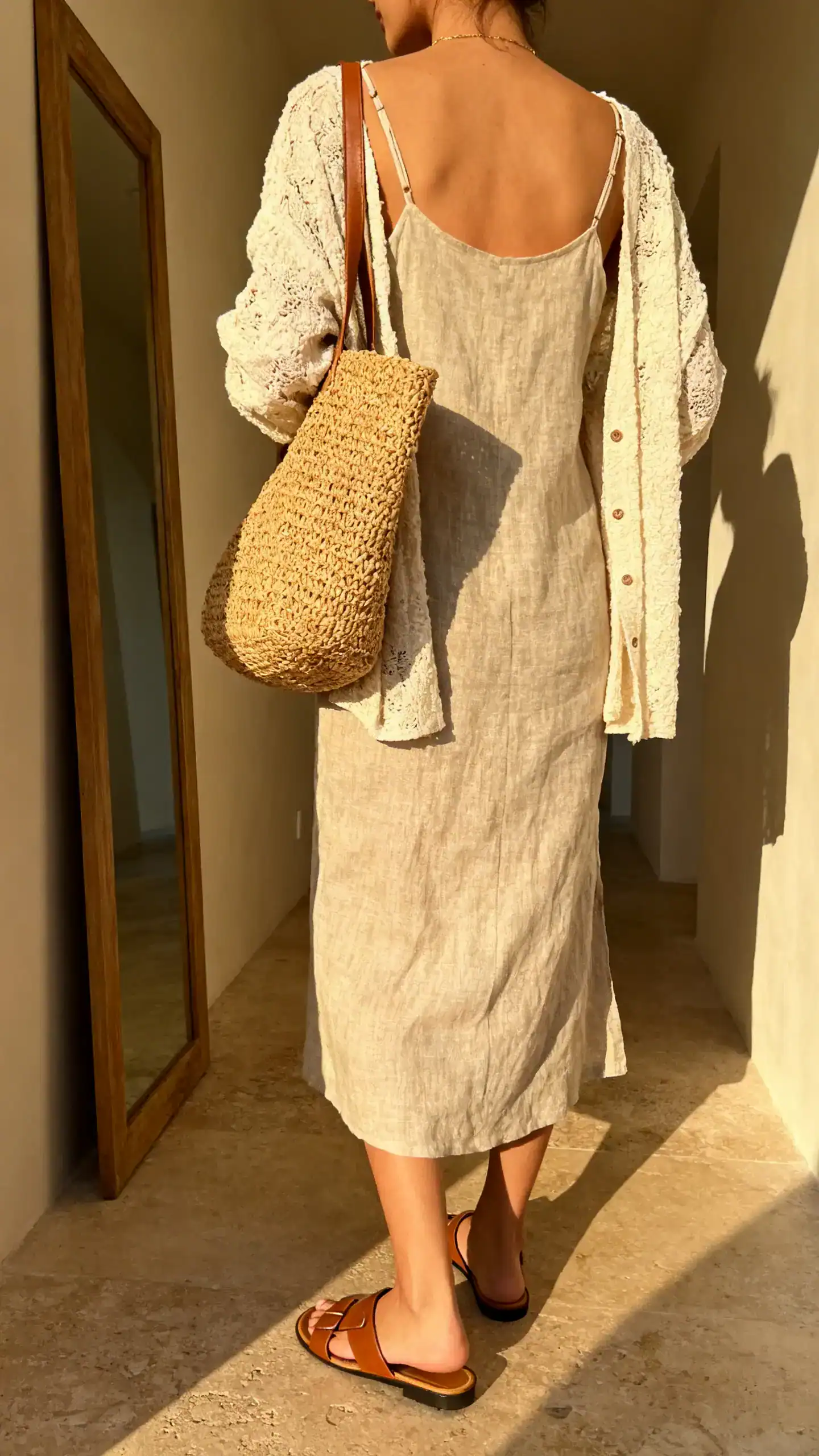 iPhone back view of a woman wearing a linen-blend slip dress layered with a lightweight coastal-texture cardigan and woven straw tote, leather slide sandals, face not visible, hallway mirror, soft afternoon light, iPhone photo quality