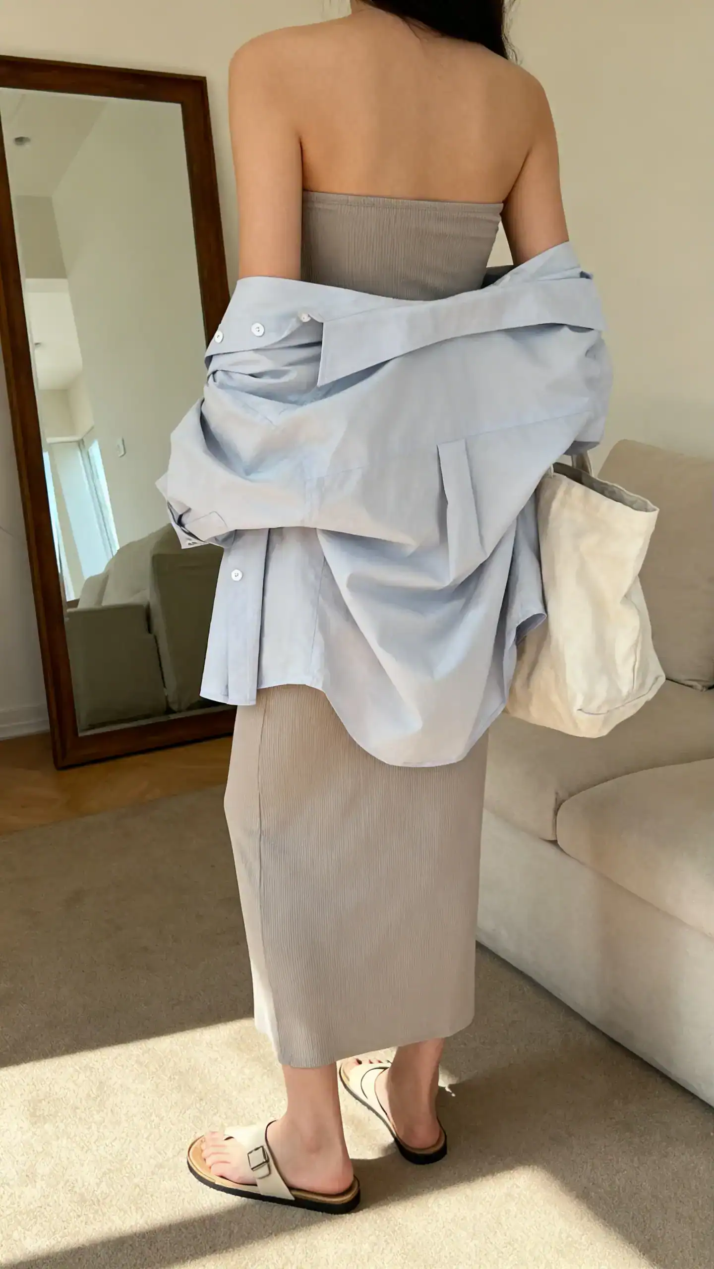 iPhone back view of a woman wearing a strapless column dress layered with an oversized button-up shirt, flat minimalist sandals, and a slouchy tote, face not visible, living room mirror, soft afternoon light, iPhone photo quality