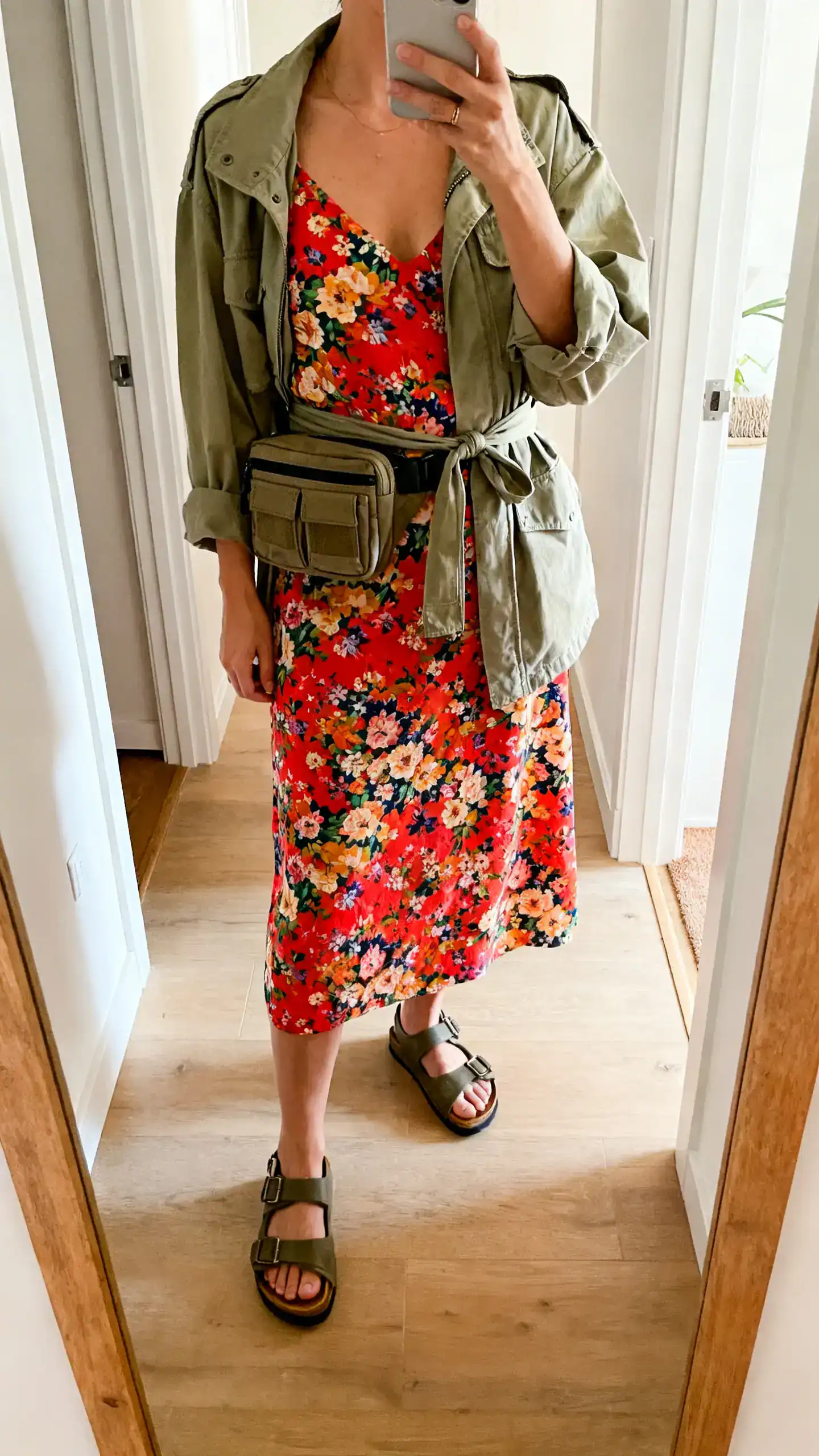 iPhone mirror selfie of a woman in a bold floral sundress with utility extras: cargo-style belt bag, lightweight field jacket tied at waist, and sturdy sandals, face not visible, cropped at neck, entryway mirror, natural daylight, iPhone photo quality