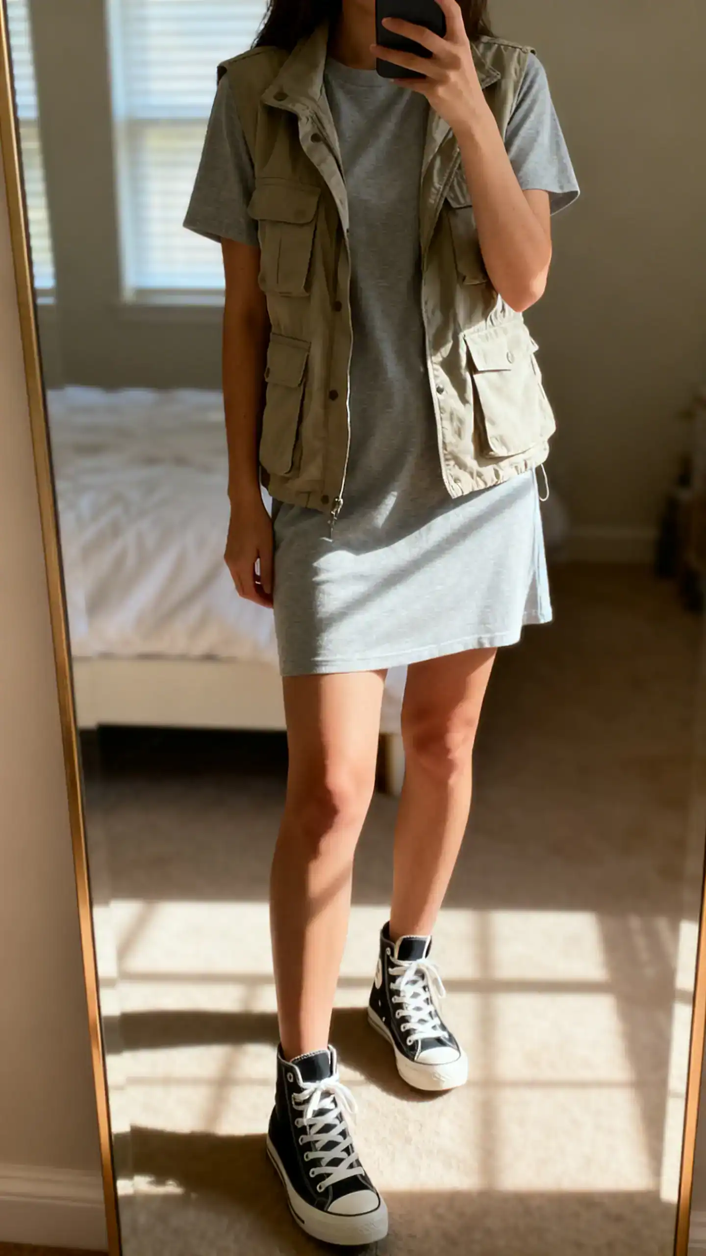 iPhone mirror selfie of a woman in a casual T-shirt dress with high-top sneakers and a lightweight utility vest, face not visible, bedroom mirror, natural window light, iPhone photo quality