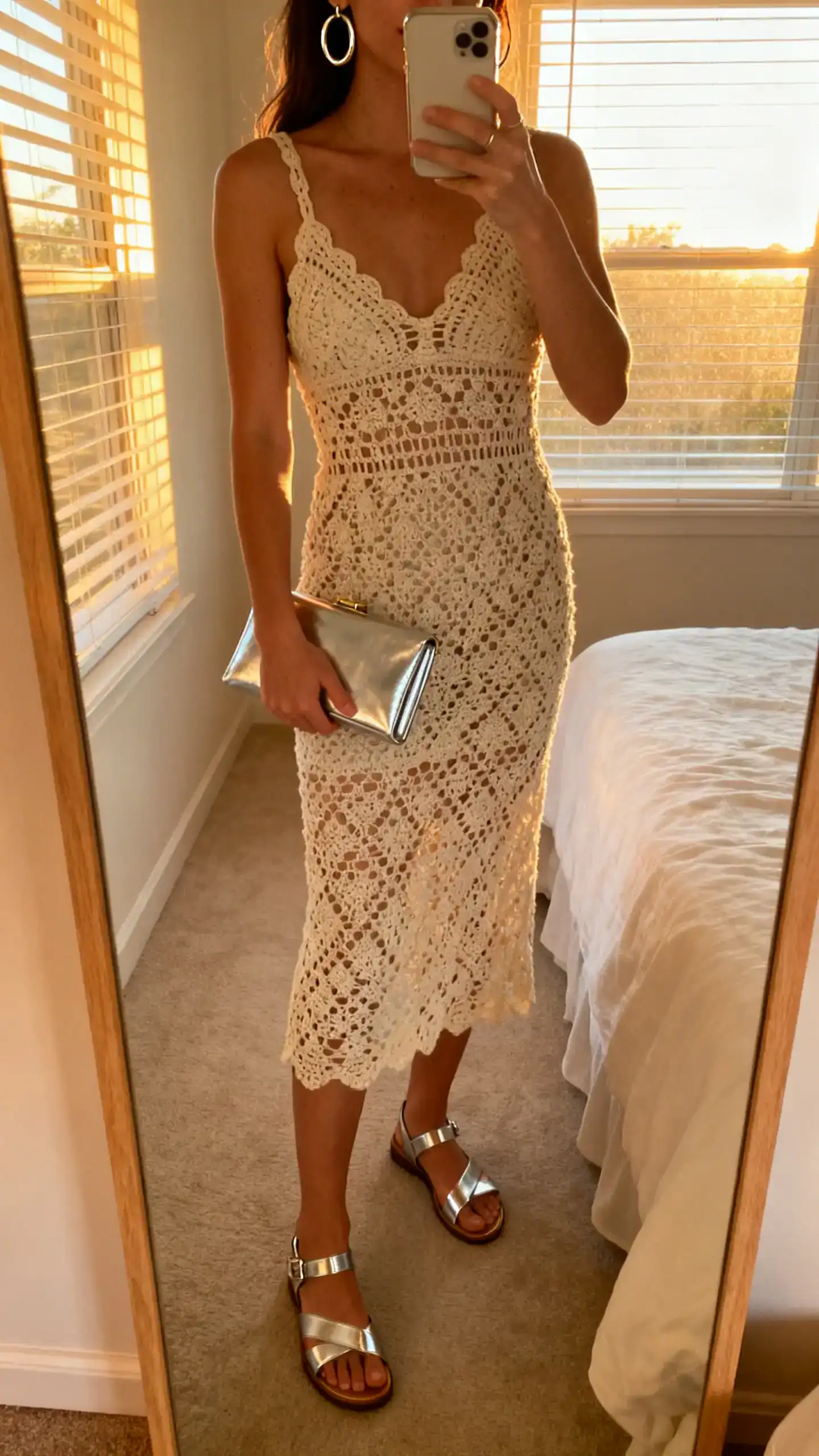 iPhone mirror selfie of a woman in a crochet midi dress accented with metallic sandals, metallic hoop earrings, and a slim metallic clutch, face not visible, cropped at neck, bedroom mirror, golden hour window light, iPhone photo quality