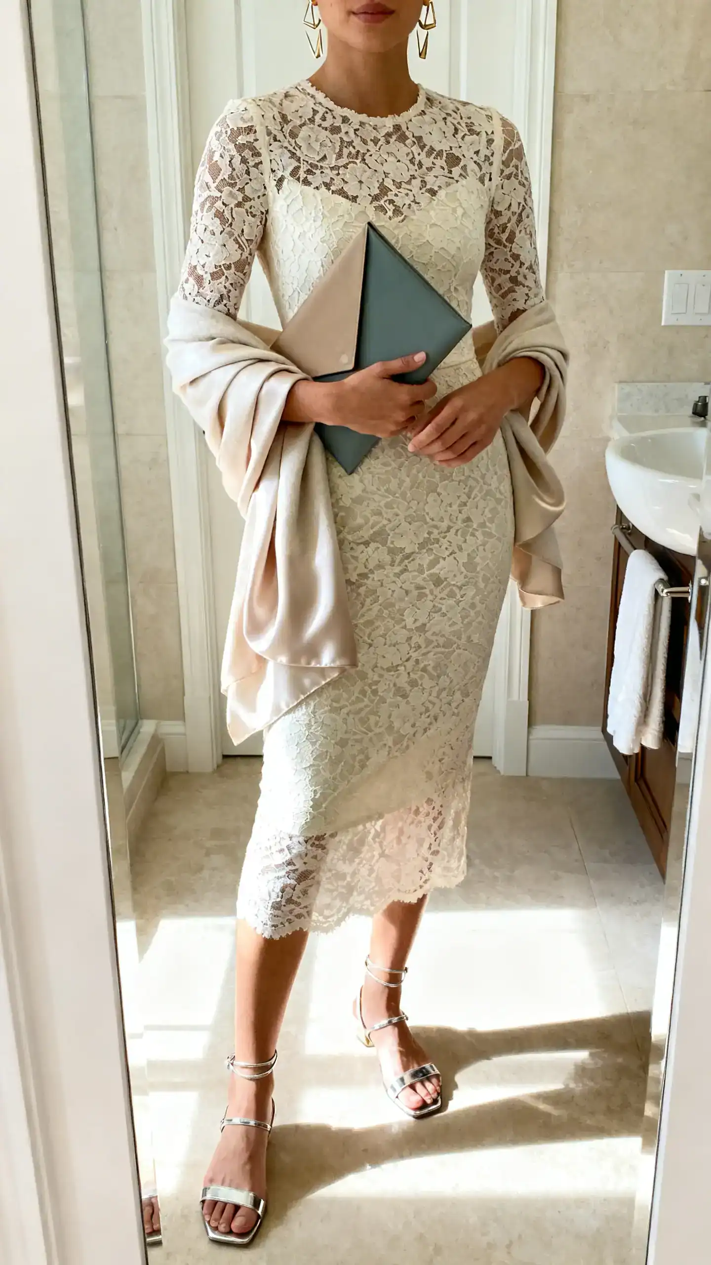 iPhone mirror selfie of a woman in a fitted lace midi sheath dress, metallic or patent barely-there sandals, ear climbers or asymmetric earrings, an envelope clutch in a contrasting shade, and a light silk/cashmere-blend wrap; face not visible, cropped at shoulders, bathroom mirror, bright indoor lighting.