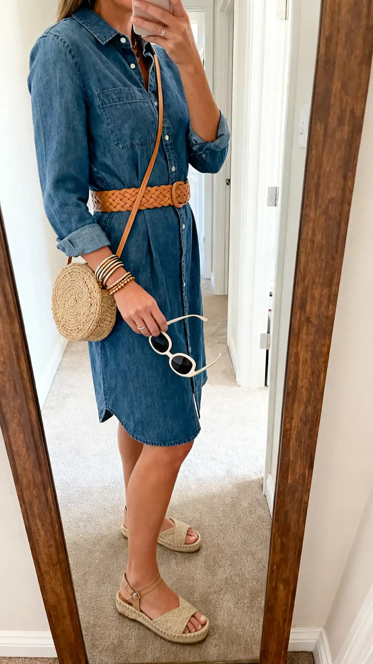 iPhone mirror selfie of a woman in a knee-length denim shirtdress with a tan woven belt, espadrille wedges, a raffia crossbody bag, stacked bangles, and oversized sunglasses in hand, face not visible, side angle in hallway mirror, morning light, iPhone photo quality.