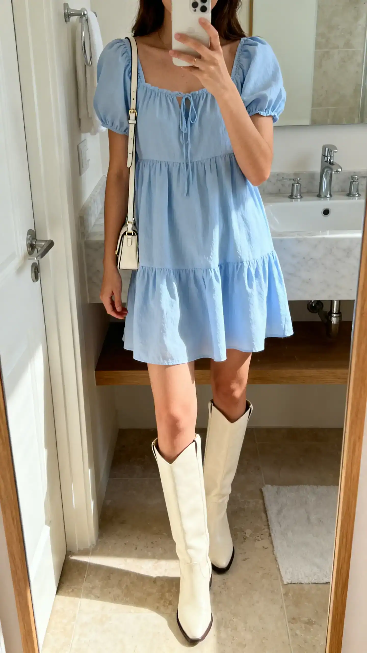 iPhone mirror selfie of a woman in a powder blue babydoll dress with cream knee-high boots and a small shoulder bag, face not visible, cropped at shoulders, bathroom mirror, bright natural light, iPhone photo quality