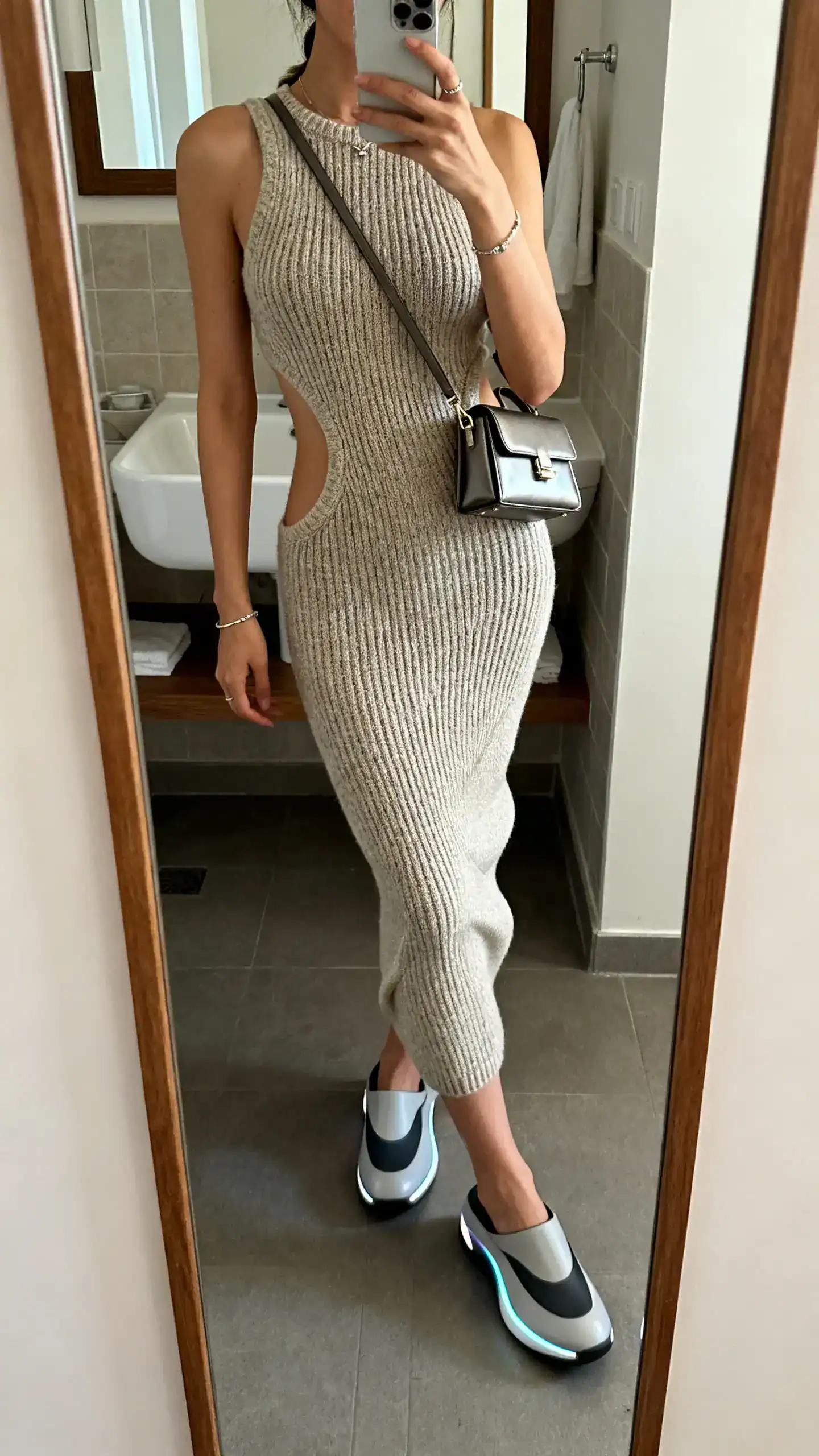 iPhone mirror selfie of a woman in a sculpted knit dress paired with futuristic flat shoes and a sleek mini shoulder bag, minimal jewelry, face not visible, bathroom mirror, indoor lighting, iPhone photo quality