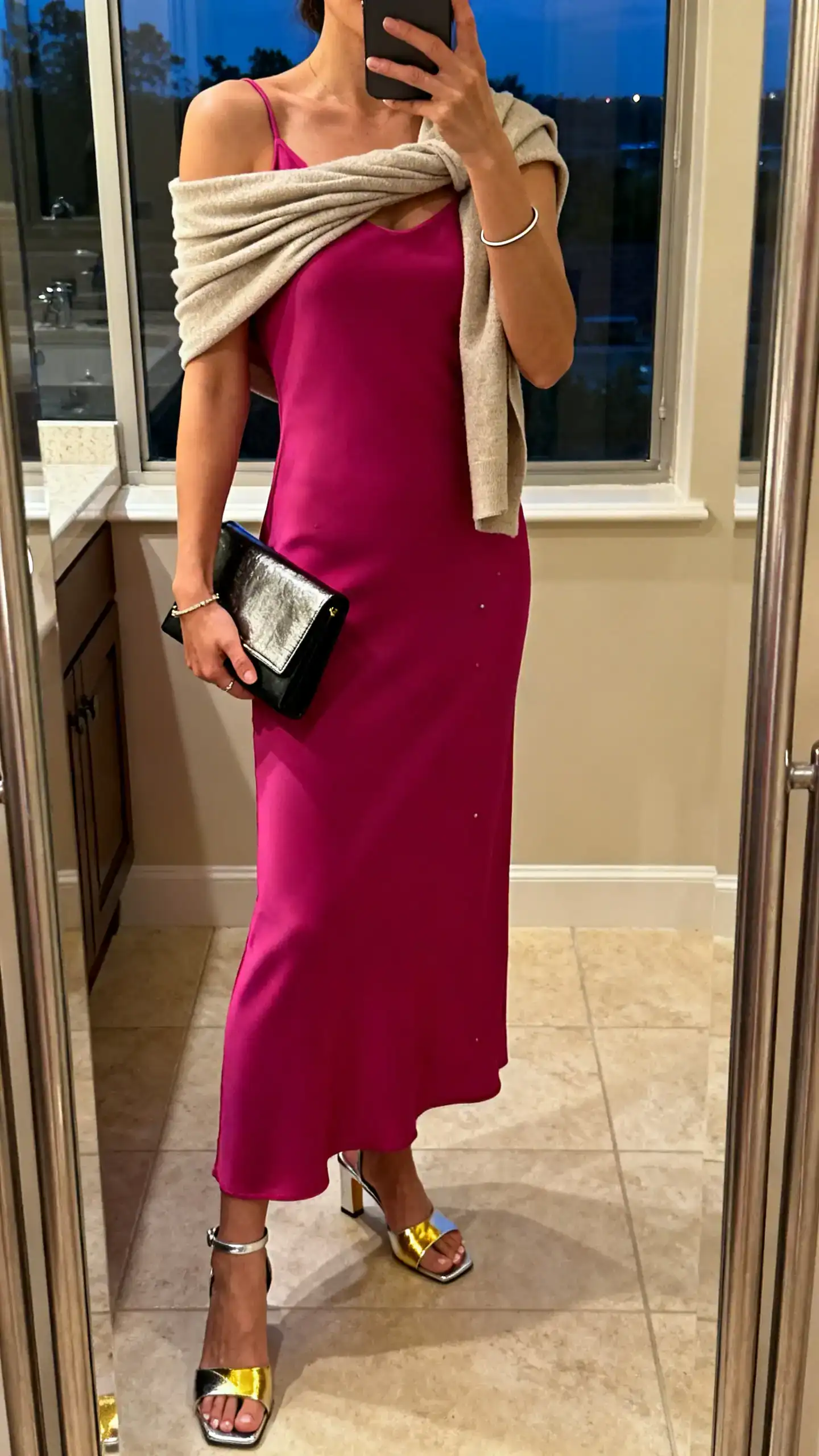 iPhone mirror selfie of a woman in a sleek column midi or slip-style maxi dress in a bold color, metallic sandals or pumps (gold/silver), a structured clutch (metallic or black), clean studs, and a thin bangle; optional cropped wrap over shoulders, face not visible, bathroom mirror, evening indoor lighting.