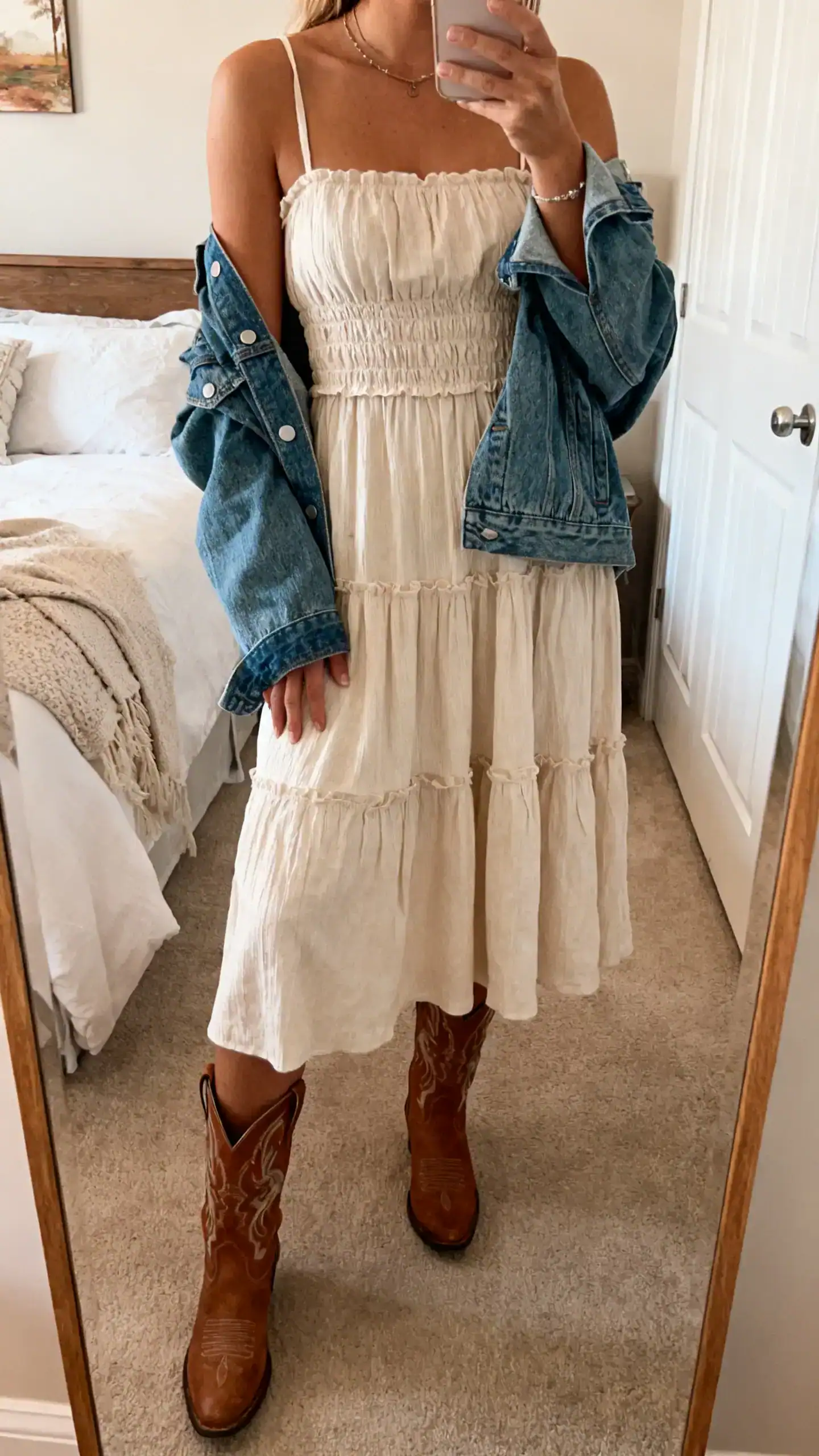 iPhone mirror selfie of a woman in a smocked midi dress with Western boots and a denim jacket draped over shoulders, festival-ready, face not visible, cropped at neck, bedroom mirror, warm indoor light, iPhone photo quality