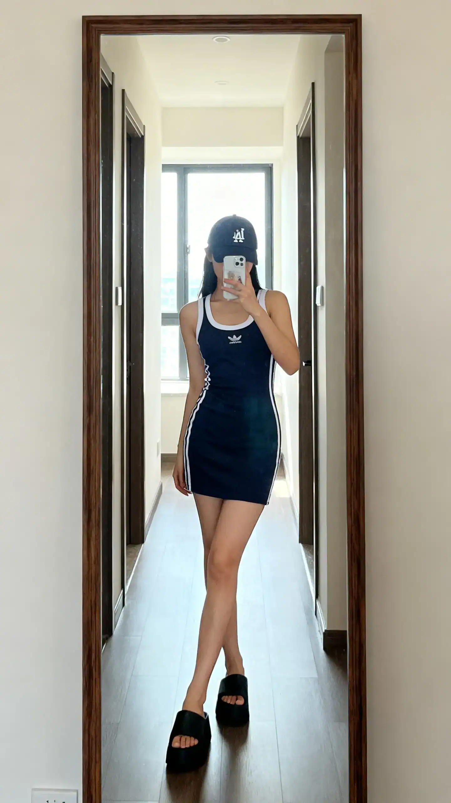 iPhone mirror selfie of a woman in a sporty tank dress styled with a baseball cap and platform slides, face not visible, cropped at shoulders, hallway full-length mirror, natural window light, iPhone photo quality