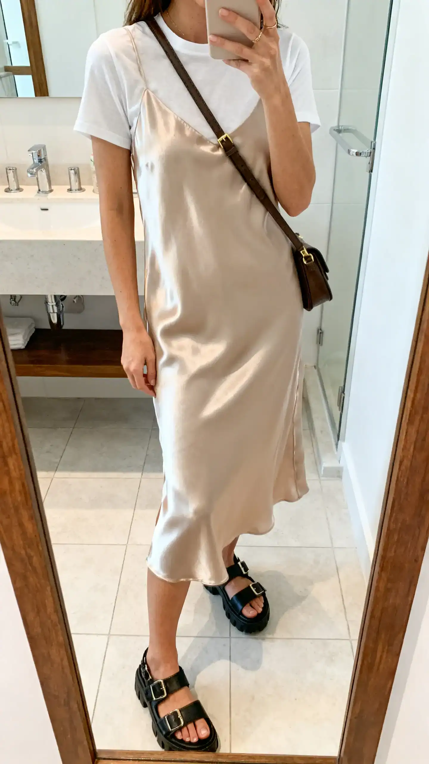 iPhone mirror selfie of a woman layering a plain tee under a satin slip dress with chunky sandals and a crossbody bag, face not visible, cropped at neck, bathroom mirror, bright indoor light, iPhone photo quality