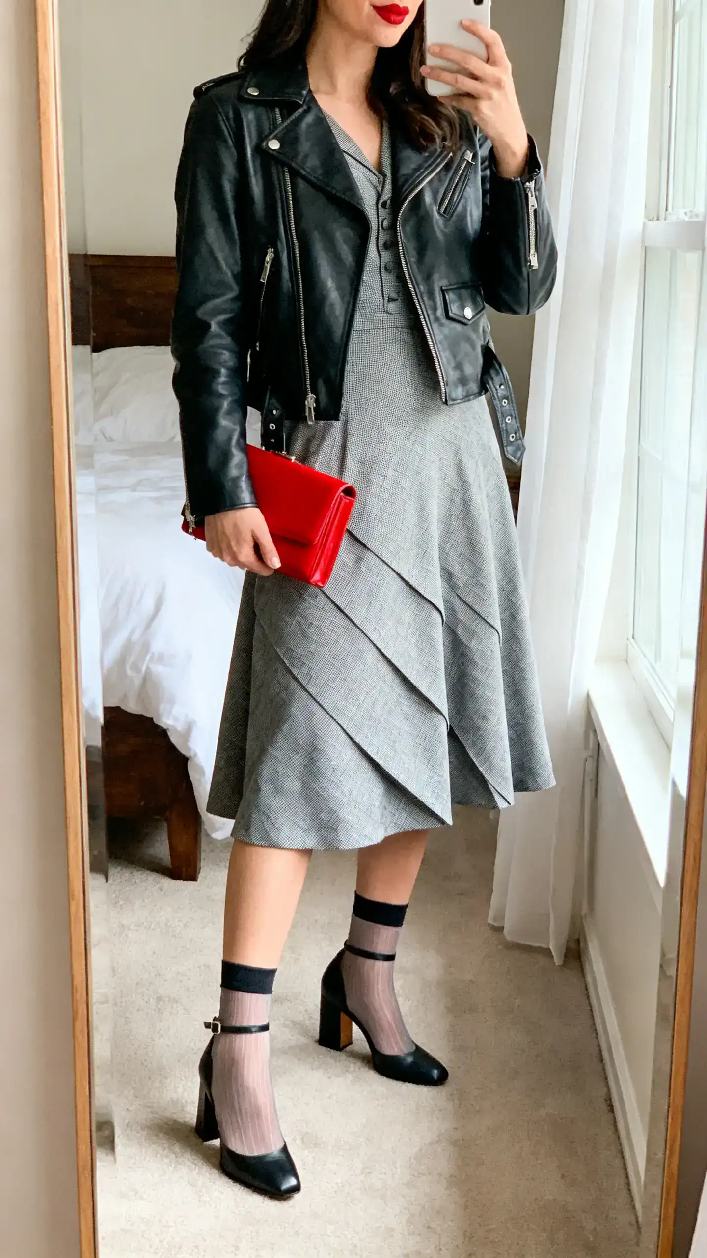 iPhone mirror selfie of a woman wearing a 1940s-inspired tea dress with a bias cut, leather moto jacket, ankle-strap block heels, sheer socks, and a red lip clutch; face not visible, cropped at shoulders, bedroom mirror, afternoon window light, iPhone photo quality.