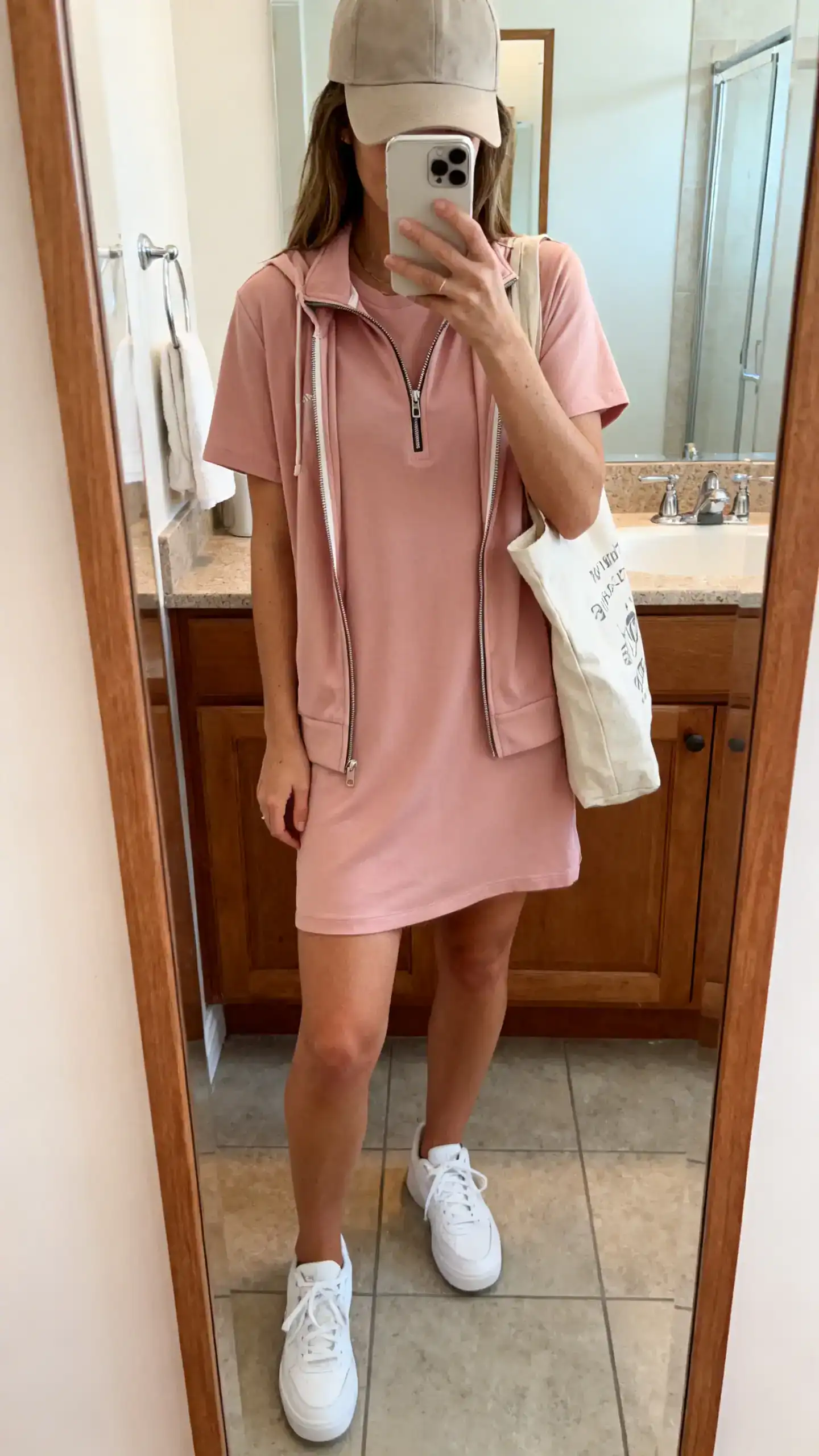 iPhone mirror selfie of a woman wearing a blush T-shirt dress with a sporty zip-up and baseball cap, white trainers and a canvas tote, face not visible, cropped at shoulders, bathroom mirror, indoor lighting, iPhone photo quality