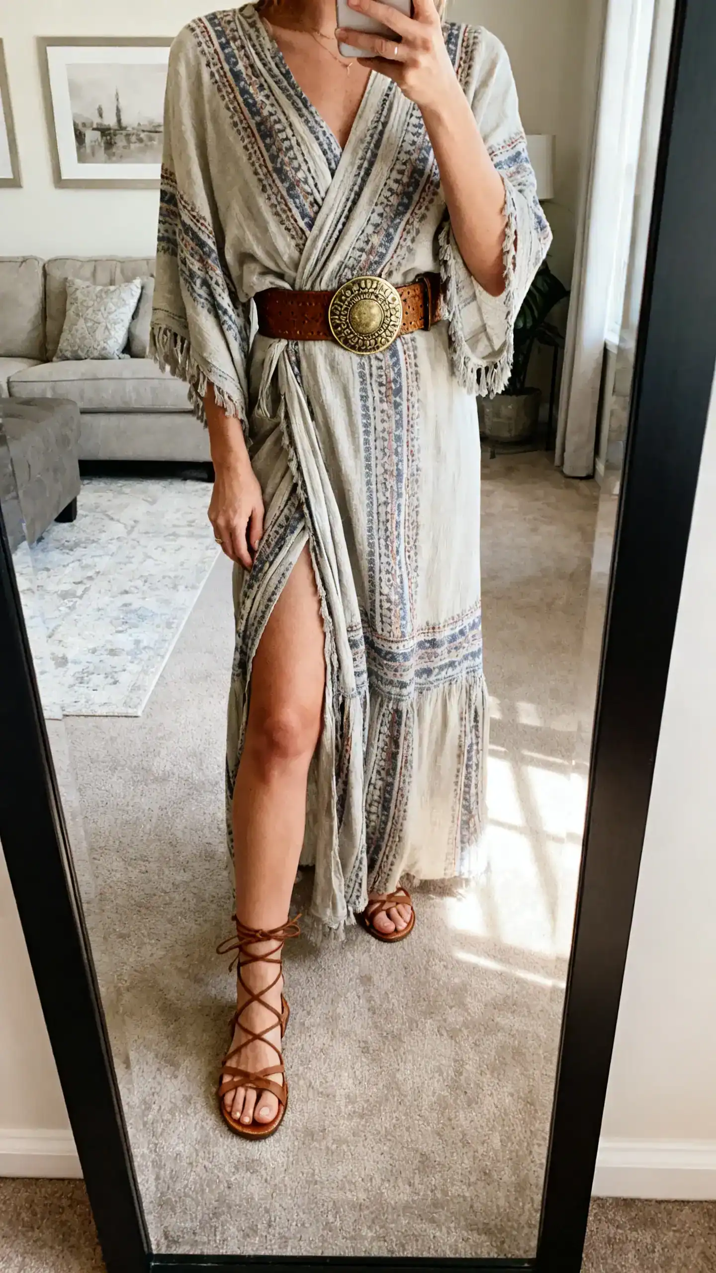 iPhone mirror selfie of a woman wearing a boho wrap maxi dress with lace-up sandals and a wide statement belt, face not visible, cropped at shoulders, living room full-length mirror, soft afternoon light, iPhone photo quality