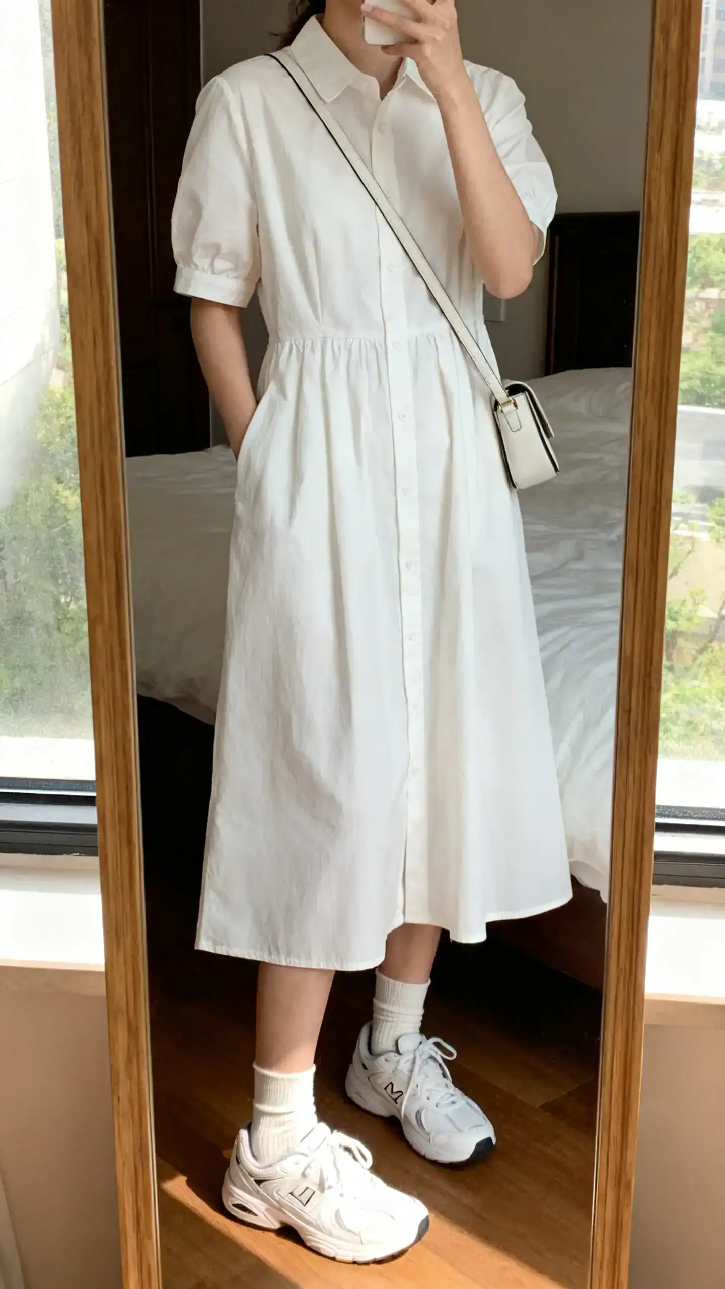 iPhone mirror selfie of a woman wearing a crisp poplin day dress styled with sporty white sneakers and ankle socks, simple crossbody bag, face not visible, cropped at neck, bedroom mirror, morning window light, iPhone photo quality