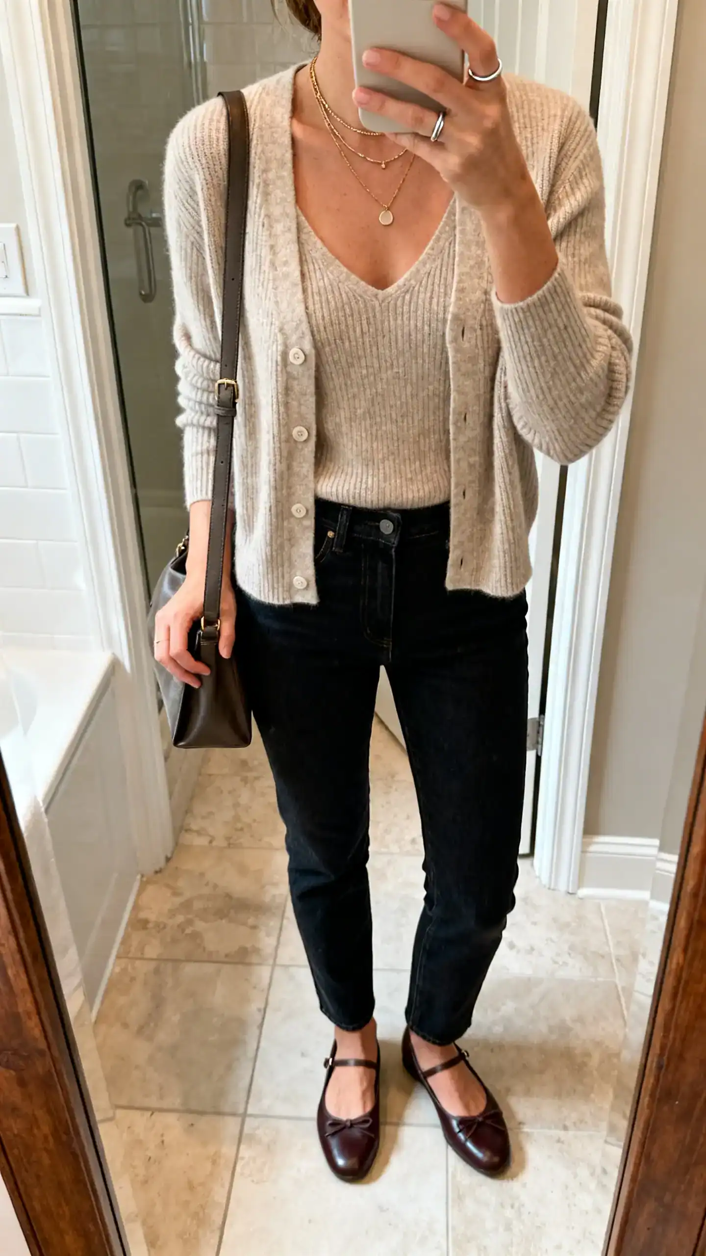 iPhone mirror selfie of a woman wearing a fine-gauge cashmere cardigan set (cardigan and matching shell) with dark slim-straight jeans, leather ballet flats, a medium-size shoulder bag, layered delicate necklaces, and a simple ring stack, face not visible, bathroom mirror, warm indoor light, iPhone photo quality.