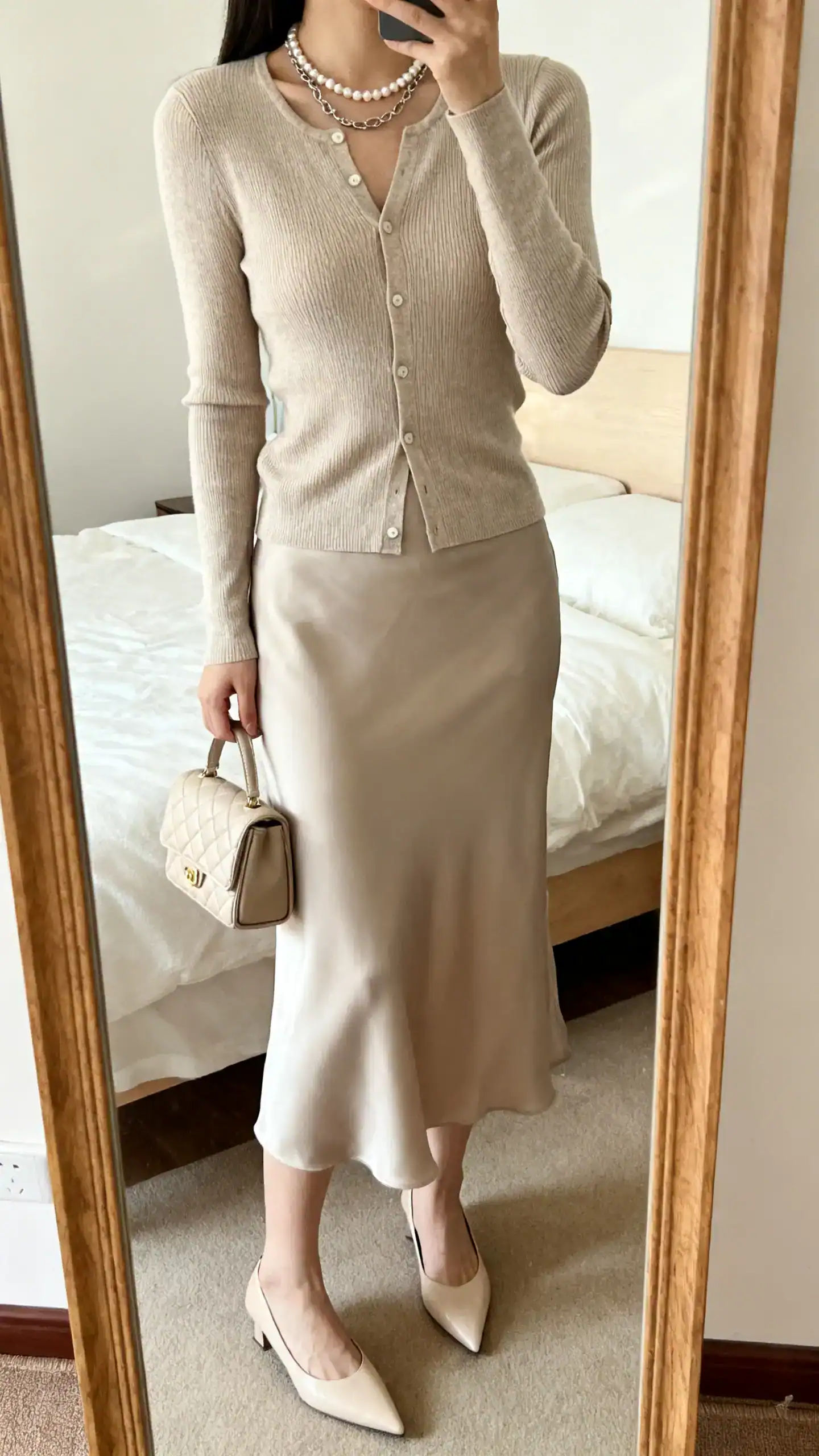 iPhone mirror selfie of a woman wearing a fitted cardigan (cashmere-silk or fine merino) with a matching or tonal slip skirt, pointed kitten heels or slingbacks, a pearl necklace or simple chain, and a quilted mini bag; face not visible, cropped at neck, bedroom mirror, soft indoor light.