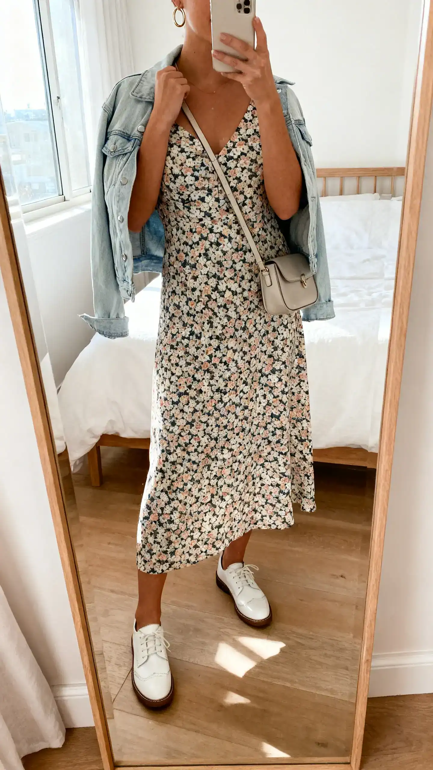iPhone mirror selfie of a woman wearing a floral midi dress with white leather sneakers, a light denim jacket draped over the shoulders, small crossbody bag, simple hoop earrings; face not visible, cropped at neck, bedroom mirror, morning window light, iPhone photo quality.
