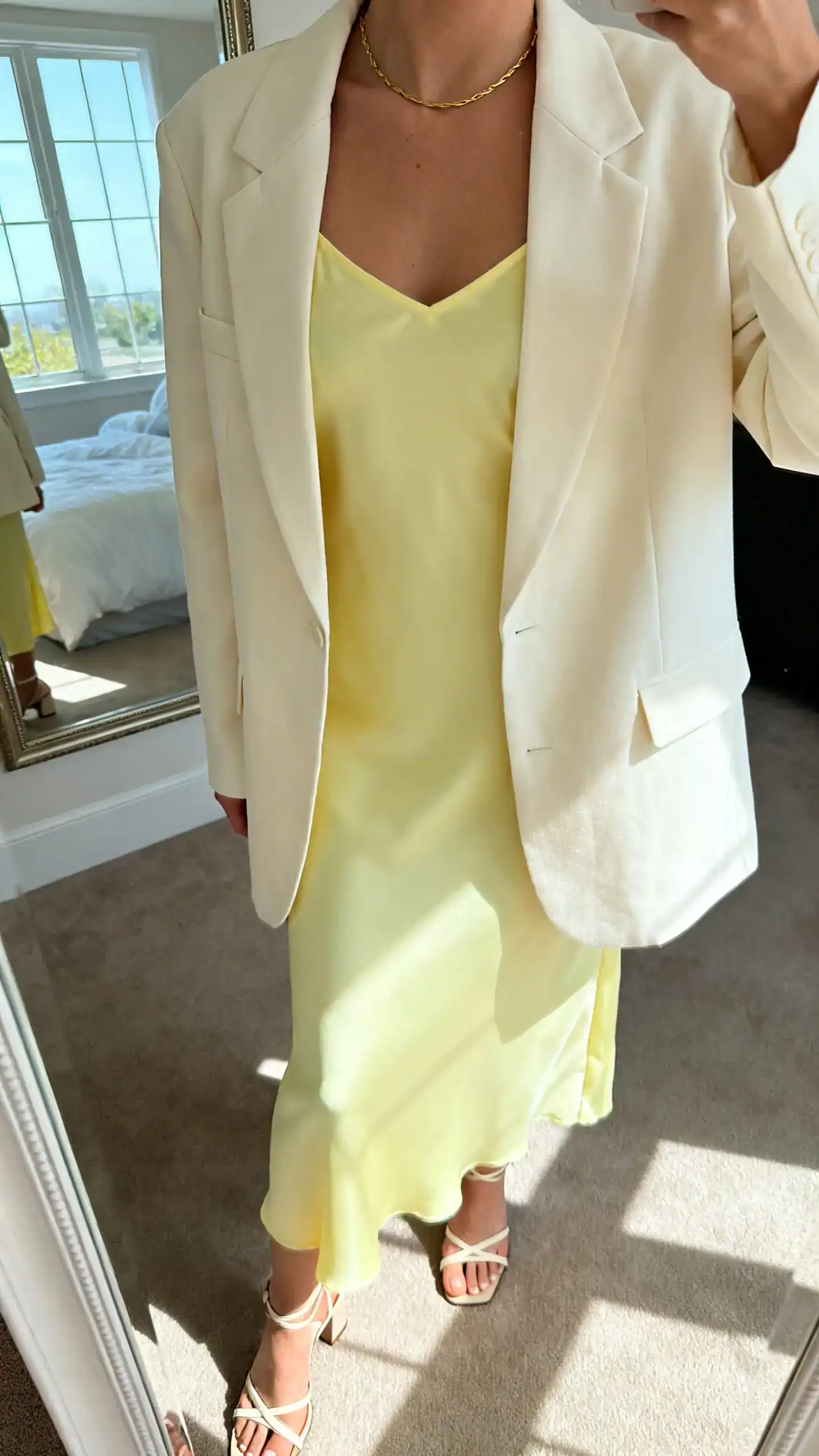 iPhone mirror selfie of a woman wearing a lemon sorbet slip dress with a tailored cream blazer, minimalist gold necklace and strappy heeled sandals, face not visible, cropped at neck, bedroom mirror, morning window light, iPhone photo quality