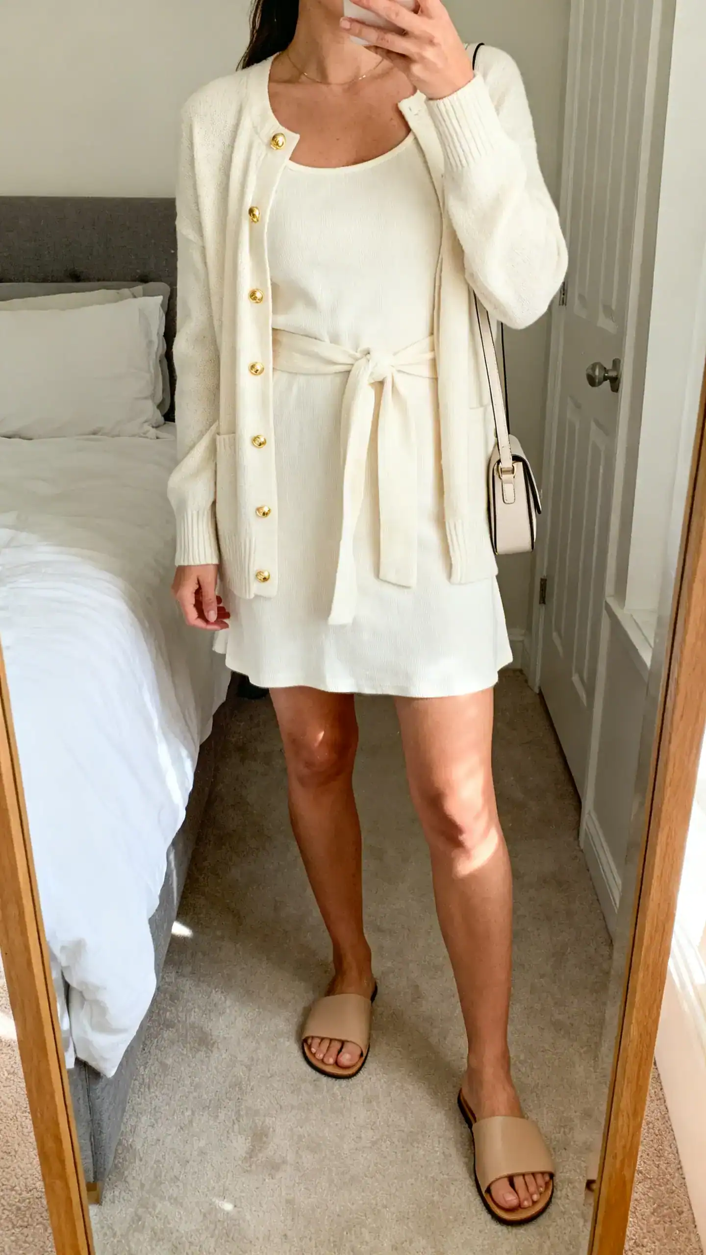 iPhone mirror selfie of a woman wearing a monochrome cream tea dress, matching cardigan tied at the waist, minimalist leather slides, slim shoulder bag, and subtle gold studs; face not visible, cropped at shoulders, bedroom mirror, soft morning light, iPhone photo quality.