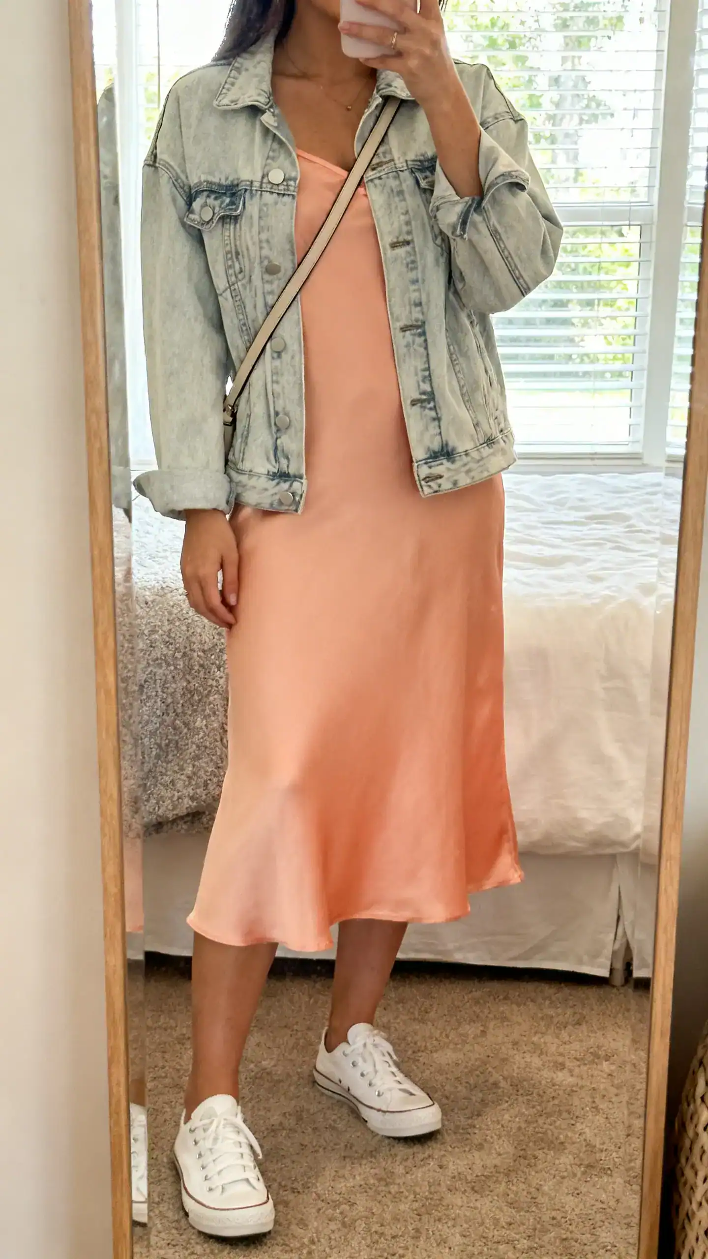 iPhone mirror selfie of a woman wearing a peach slip midi dress with a light-wash denim jacket, white sneakers and a crossbody bag, face not visible, bedroom mirror, soft afternoon window light, iPhone photo quality