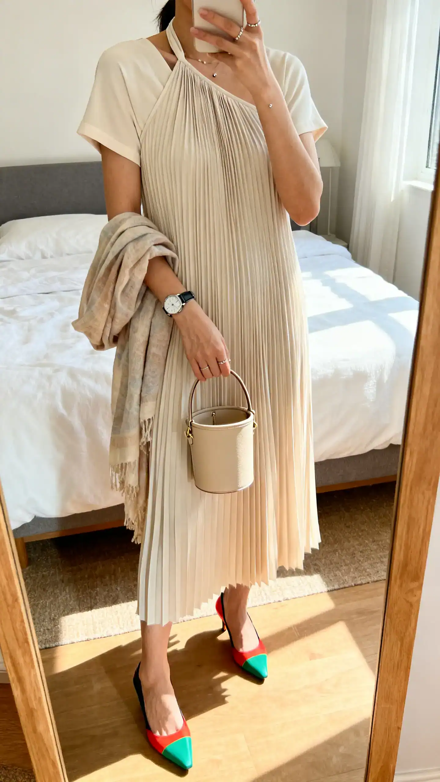 iPhone mirror selfie of a woman wearing a pleated midi/tea-length dress (halter/one-shoulder/cap-sleeve), color-pop heels, minimal studs, a sleek watch, and a compact bucket bag or top-handle mini; lightweight shawl in hand, face not visible, bedroom mirror, soft morning light, iPhone photo quality.