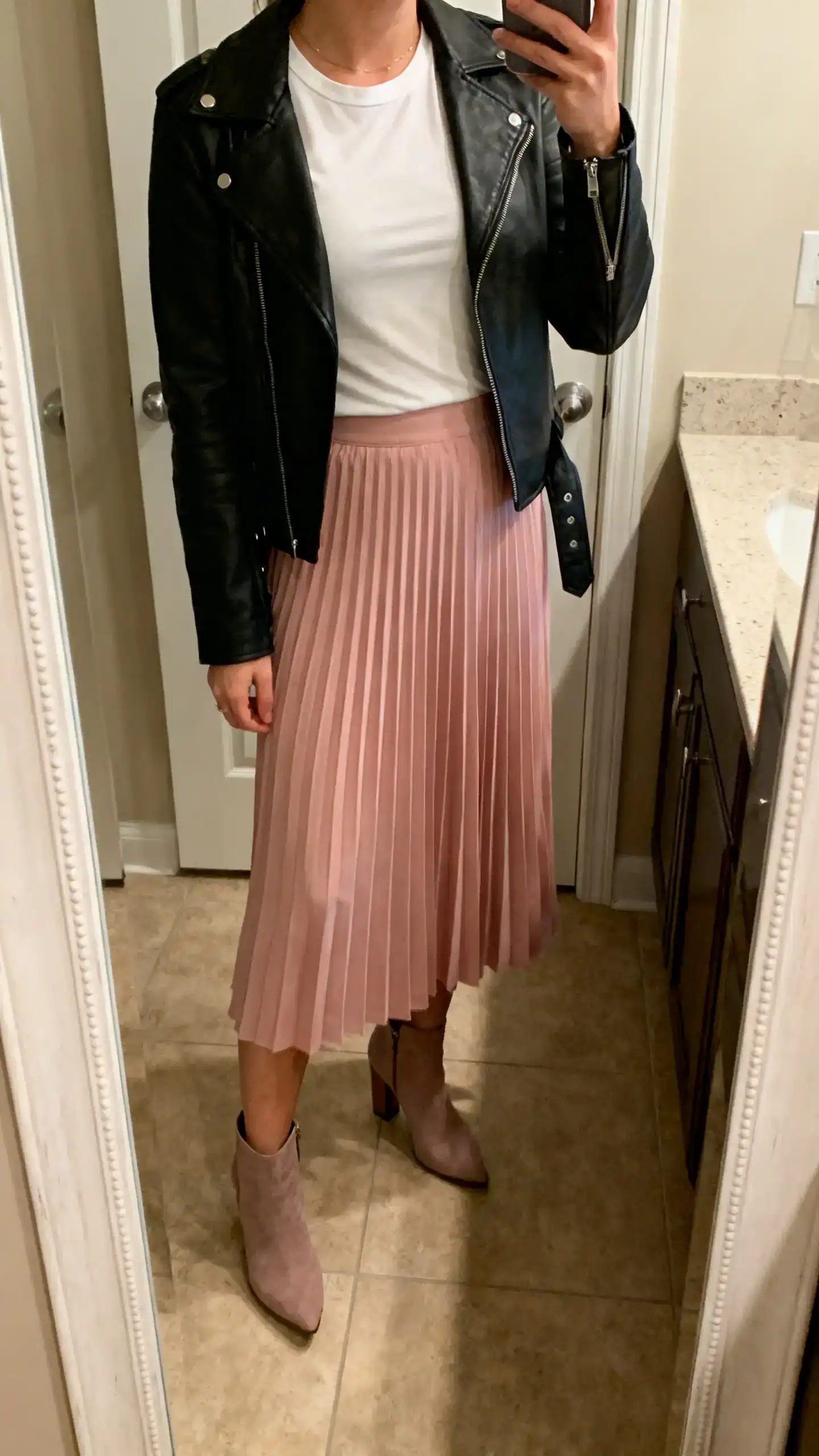 iPhone mirror selfie of a woman wearing a powder pink pleated midi skirt with a black leather jacket, fitted white tee and heeled ankle boots, face not visible, cropped at neck, bathroom mirror, warm indoor light, iPhone photo quality