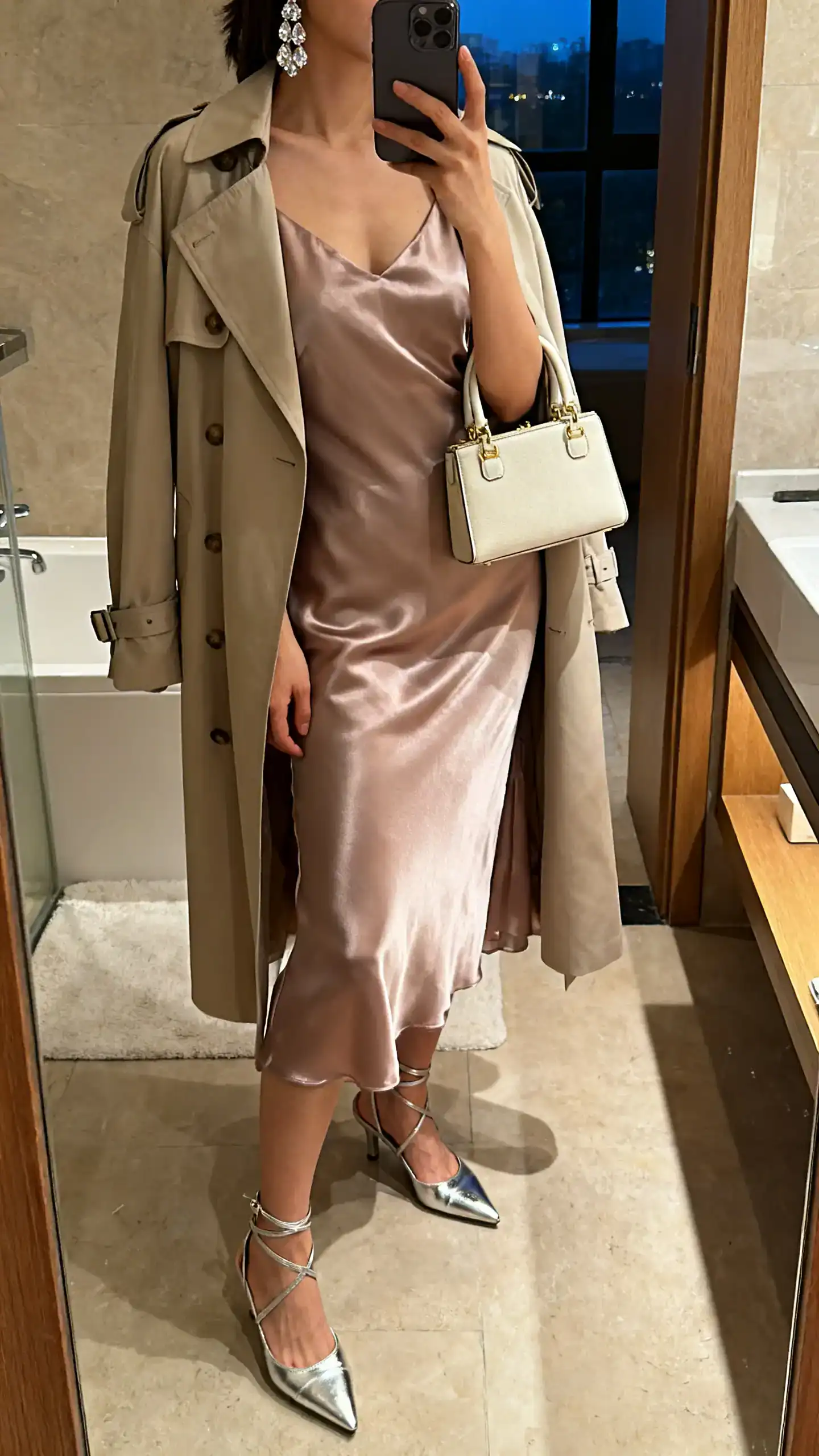 iPhone mirror selfie of a woman wearing a satin tea dress with a lightweight trench, strappy metallic heels, crystal drop earrings, and a mini top-handle bag; face not visible, cropped at neck, bathroom mirror, warm evening indoor lighting, iPhone photo quality.