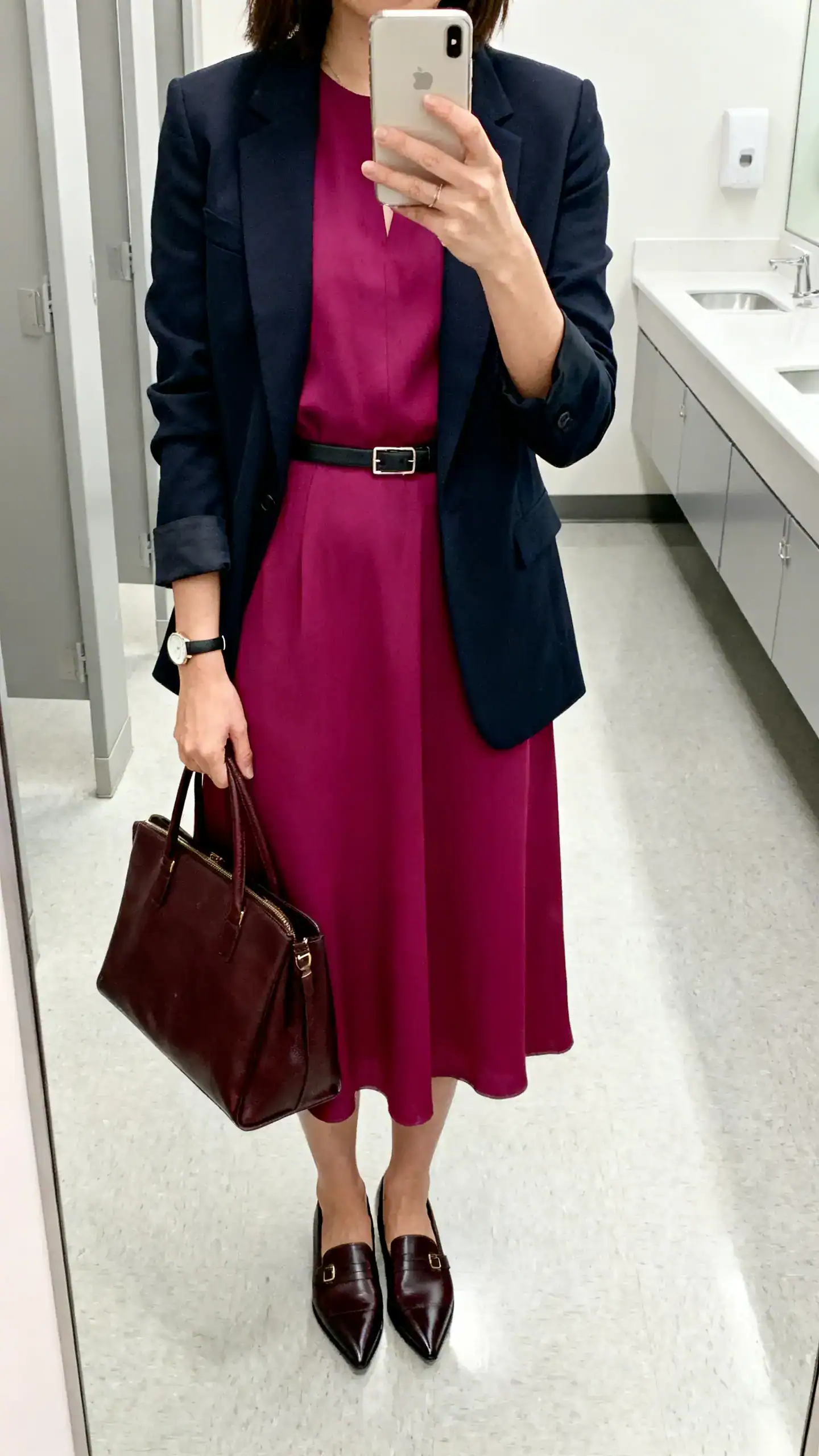 iPhone mirror selfie of a woman wearing a solid jewel-tone tea dress with a tailored blazer, pointed-toe loafers, structured leather tote, and a slim waist belt plus minimalist watch; face not visible, cropped at neck, office restroom mirror, bright indoor lighting, iPhone photo quality.