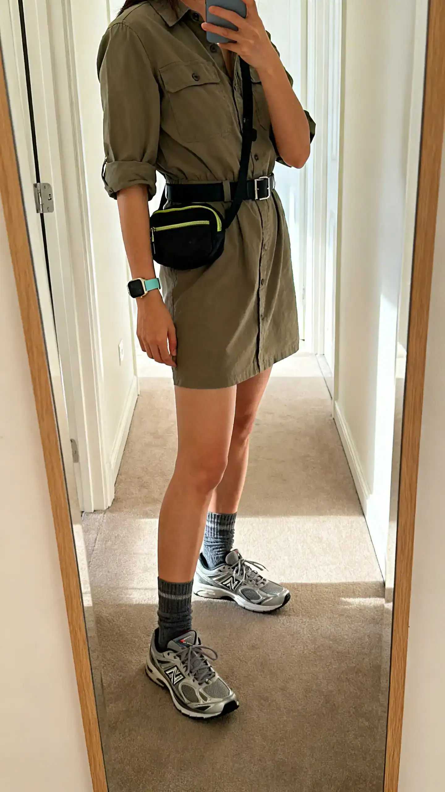 iPhone mirror selfie of a woman wearing a utility shirtwaist dress with runner sneakers, belt bag worn crossbody, sporty watch, crew socks; face not visible, side angle, hallway full-length mirror, indoor daylight, iPhone photo quality.