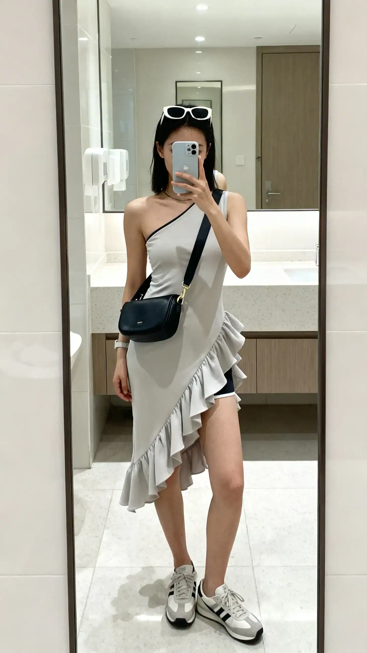 iPhone mirror selfie of a woman wearing an asymmetric ruffle dress with sleek sport accessories: streamlined crossbody, sporty sunglasses, and minimalist trainers, face not visible, bathroom mirror, bright indoor lighting, iPhone photo quality