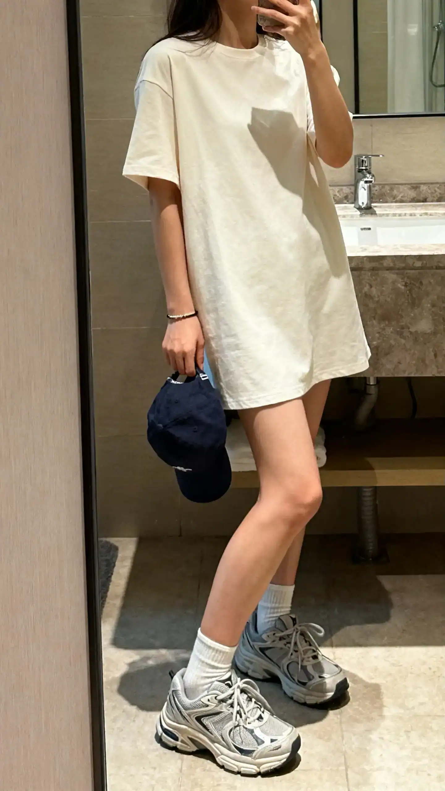 iPhone side-angle mirror selfie of a woman in a cotton T-shirt dress with chunky dad sneakers, baseball cap in hand, ankle socks, minimal bracelet; face not visible, shoulder-crop, bathroom mirror, soft indoor lighting, iPhone photo quality.