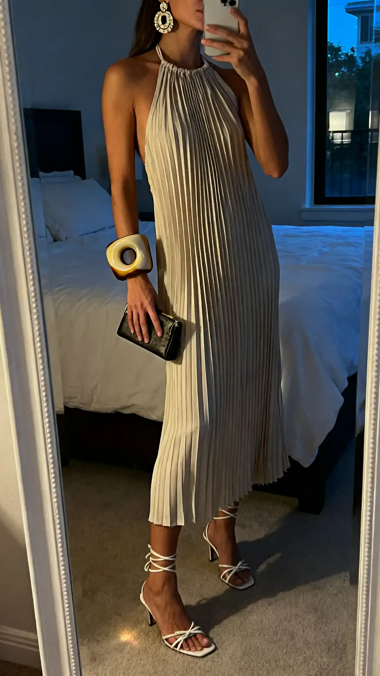 iPhone side-angle mirror selfie of a woman in a pleated halter dress with sculptural jewelry (bold cuff and statement earrings) and strappy heels, small clutch, face not visible, bedroom mirror, evening indoor light, iPhone photo quality