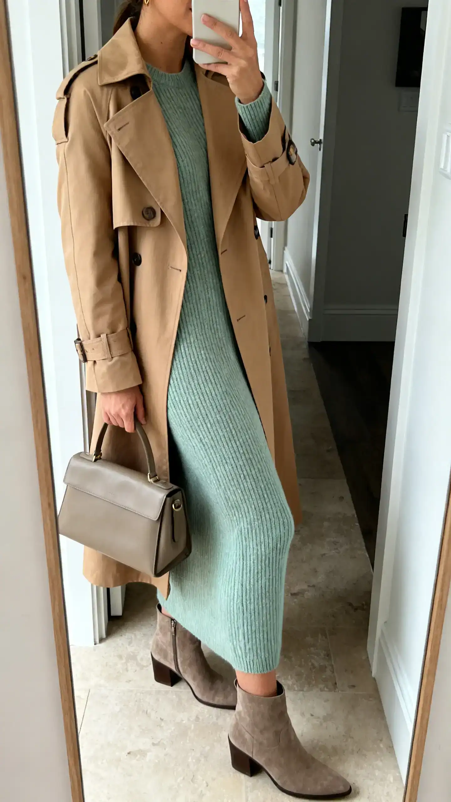 iPhone side-angle mirror selfie of a woman in a soft sage knit dress layered with a tan trench coat, ankle boots and a structured tote, face not visible, entryway mirror, overcast natural light, iPhone photo quality