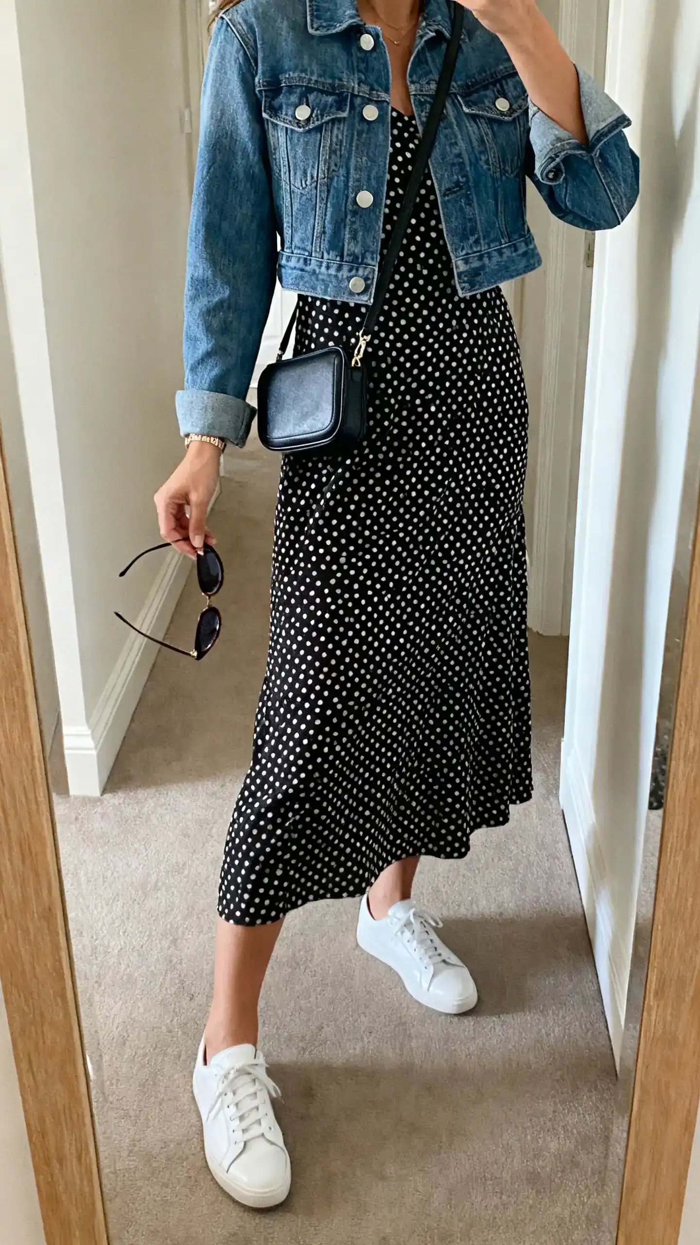 iPhone side-angle mirror selfie of a woman wearing a black-and-white polka-dot tea dress, white leather sneakers, cropped denim jacket, crossbody mini bag, and slim cat-eye sunglasses in hand; face not visible, cropped at shoulders, hallway full-length mirror, soft indoor daylight, iPhone photo quality.