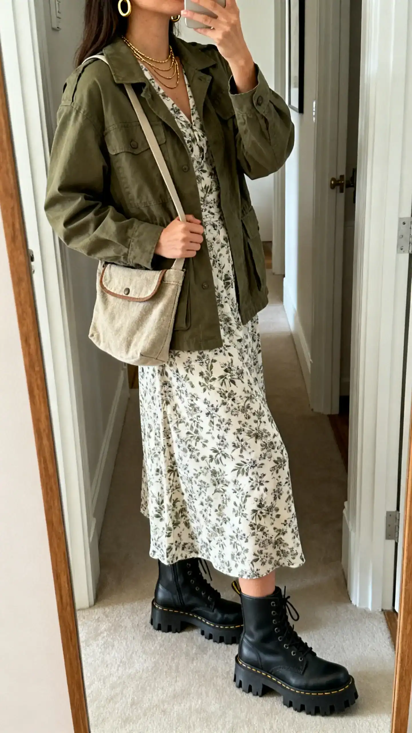 iPhone side-angle mirror selfie of a woman wearing a printed tea dress paired with an olive utility jacket, chunky lug-sole boots, canvas crossbody, and layered gold hoops; face not visible, cropped at neck, entryway full-length mirror, natural indoor light, iPhone photo quality.