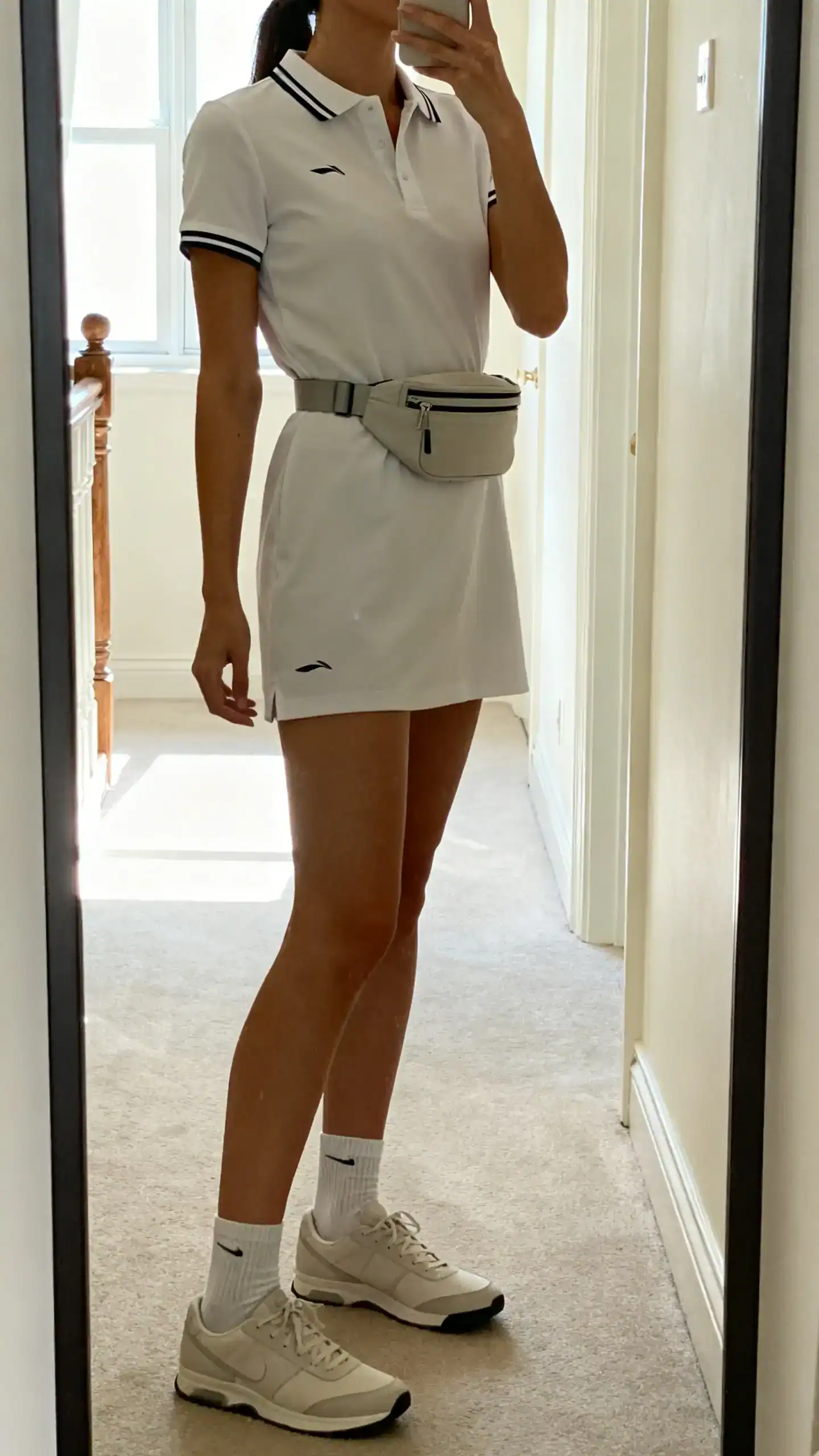 iPhone side-angle mirror selfie of a woman wearing a tennis-inspired polo dress with sleek minimal sneakers, low socks, and a streamlined belt bag, face not visible, full-length mirror in hallway, bright morning light, iPhone photo quality