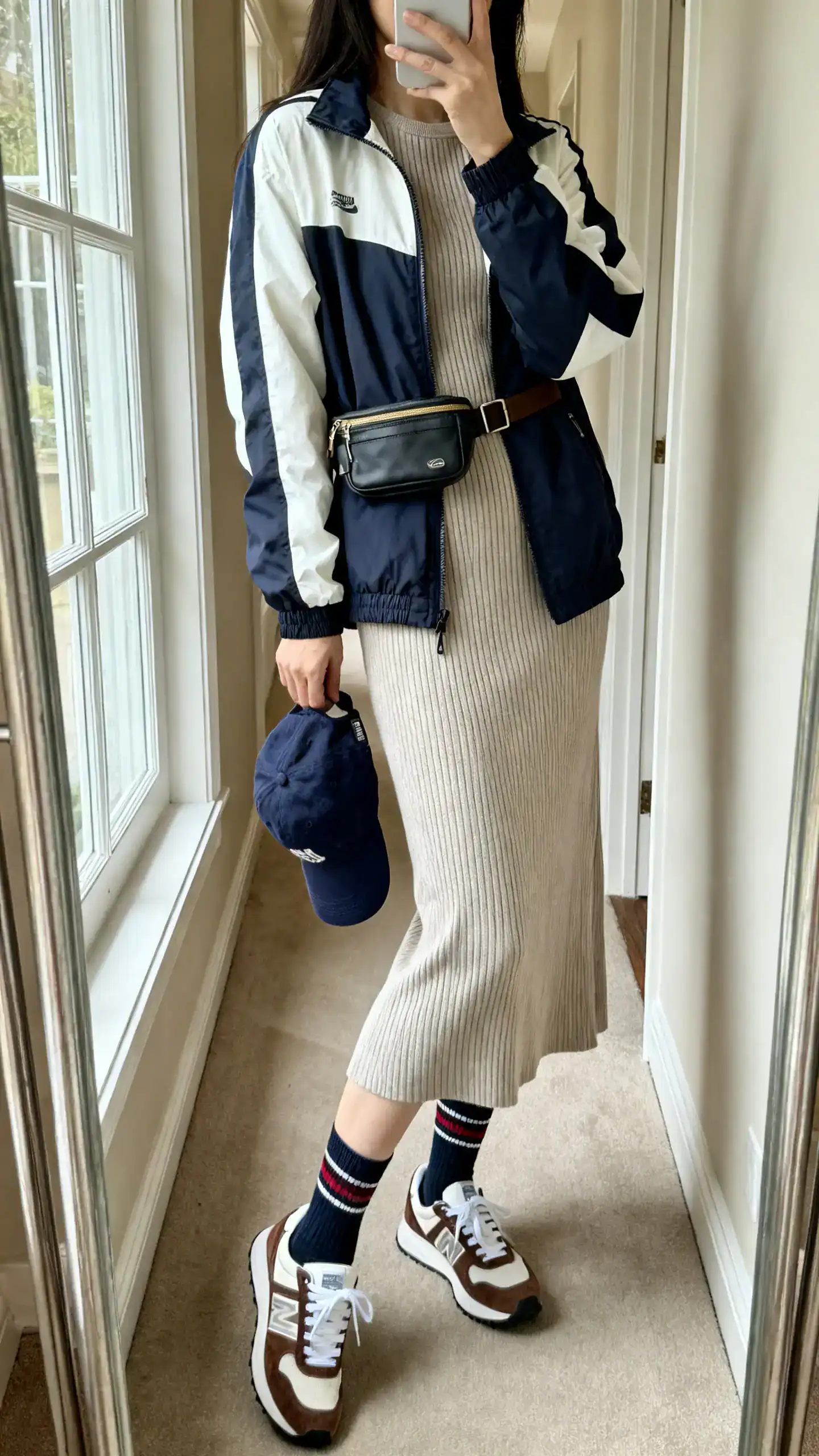 iPhone side-angle mirror selfie of a woman wearing a sporty windbreaker over a ribbed tea dress, crew socks with retro sneakers, baseball cap in hand, and a belt bag; face not visible, hallway mirror, natural window light, iPhone photo quality.