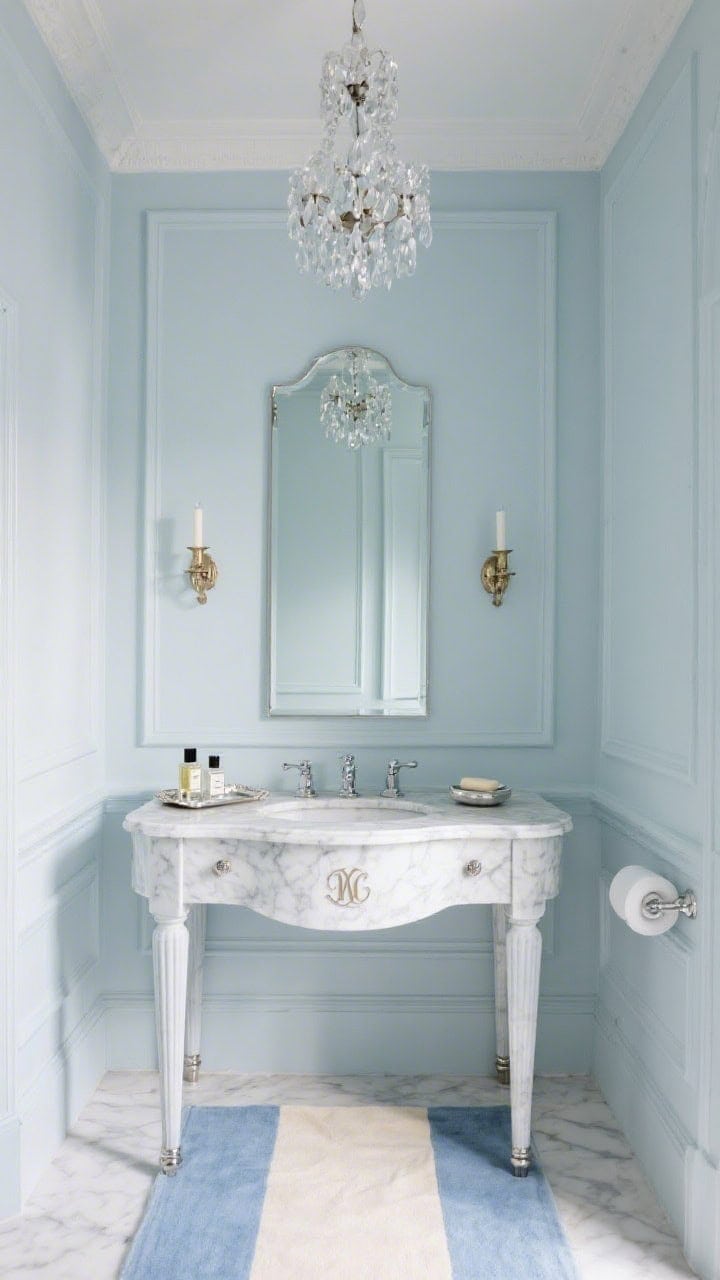 Medium shot, elegant front view: A petite palace-style bathroom with powder blue walls and refined panel mouldings. A marble-topped vanity with fluted legs and polished nickel hardware stands beneath a tall bevel-edged mirror, flanked by candle-style sconces. Overhead hangs a petite crystal chandelier; a soft blue-and-ivory runner on the floor. Add a silver tray with cologne bottles and a monogrammed soap dish. Gentle, sparkling light.
