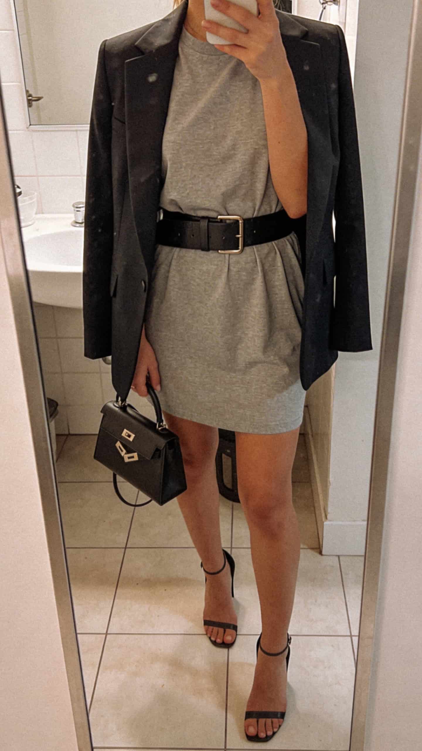 iPhone mirror selfie of a woman wearing a tailored T-shirt dress elevated with a sharp blazer, heeled sandals, structured belt, and a top-handle bag, face not visible, cropped at neck, bathroom mirror, indoor lighting, iPhone photo quality