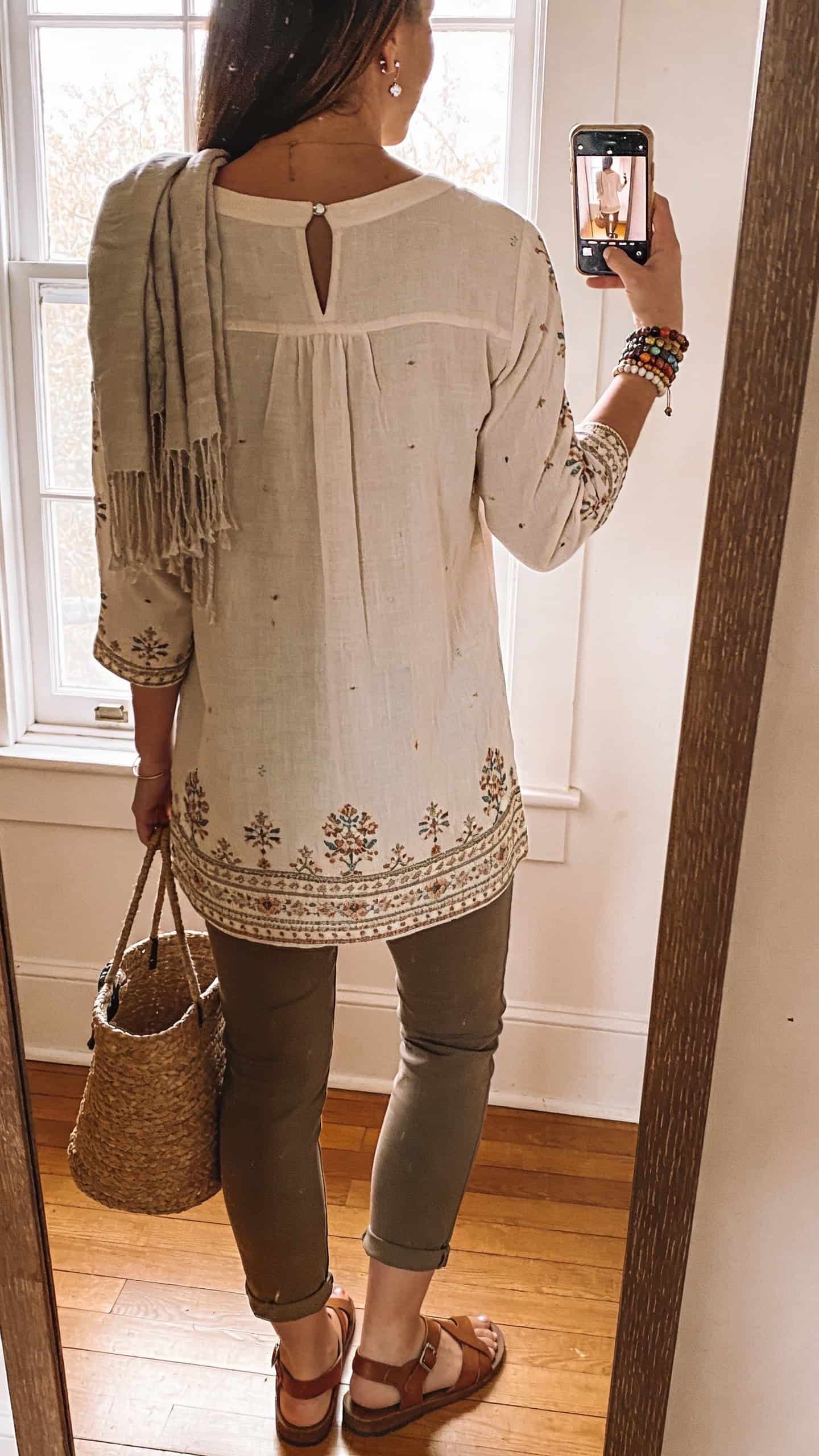 iPhone back-view mirror selfie of a woman in an embroidered cotton tunic with notched neckline and 3/4 sleeves, slim cropped pants, leather sandals, a woven bucket bag, beaded bracelet stack and delicate studs, lightweight scarf draped over shoulder, face not visible, full-length mirror, natural window light, iPhone photo quality.