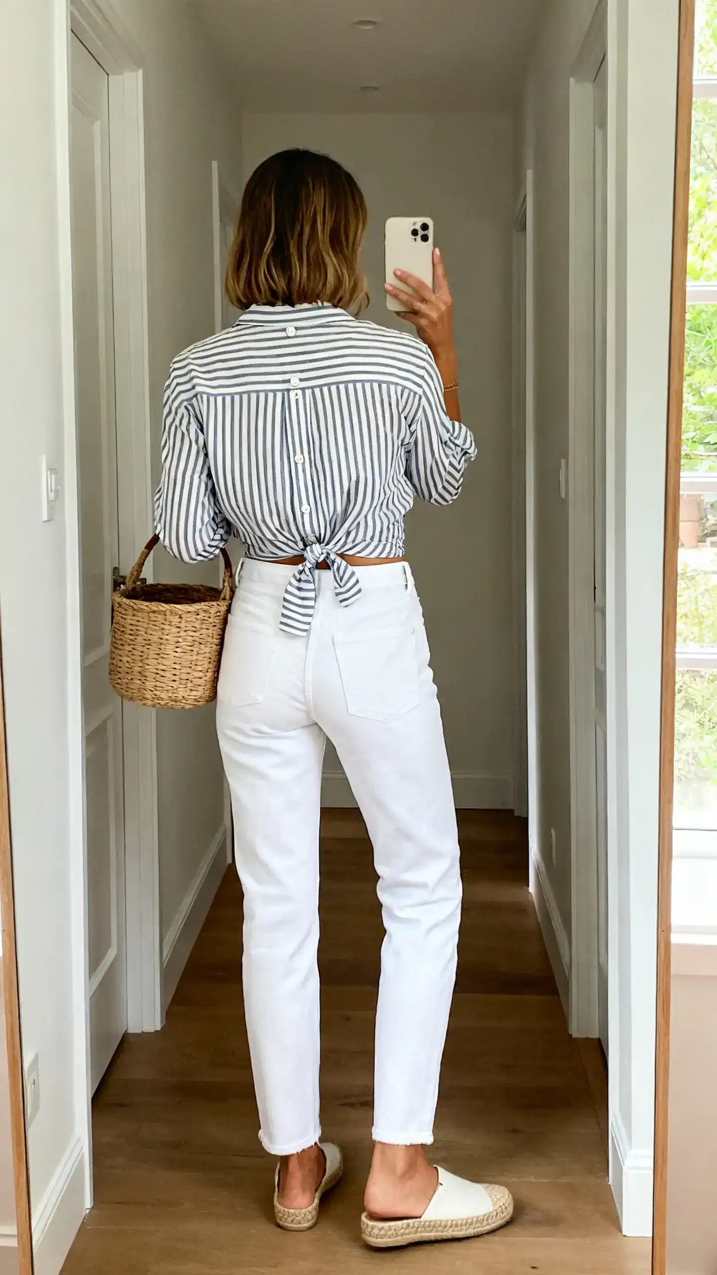 iPhone back-view hallway mirror selfie of a woman in a Riviera-striped button-up half-tucked into white jeans with espadrilles and a basket bag, face not visible, natural daylight, iPhone photo quality