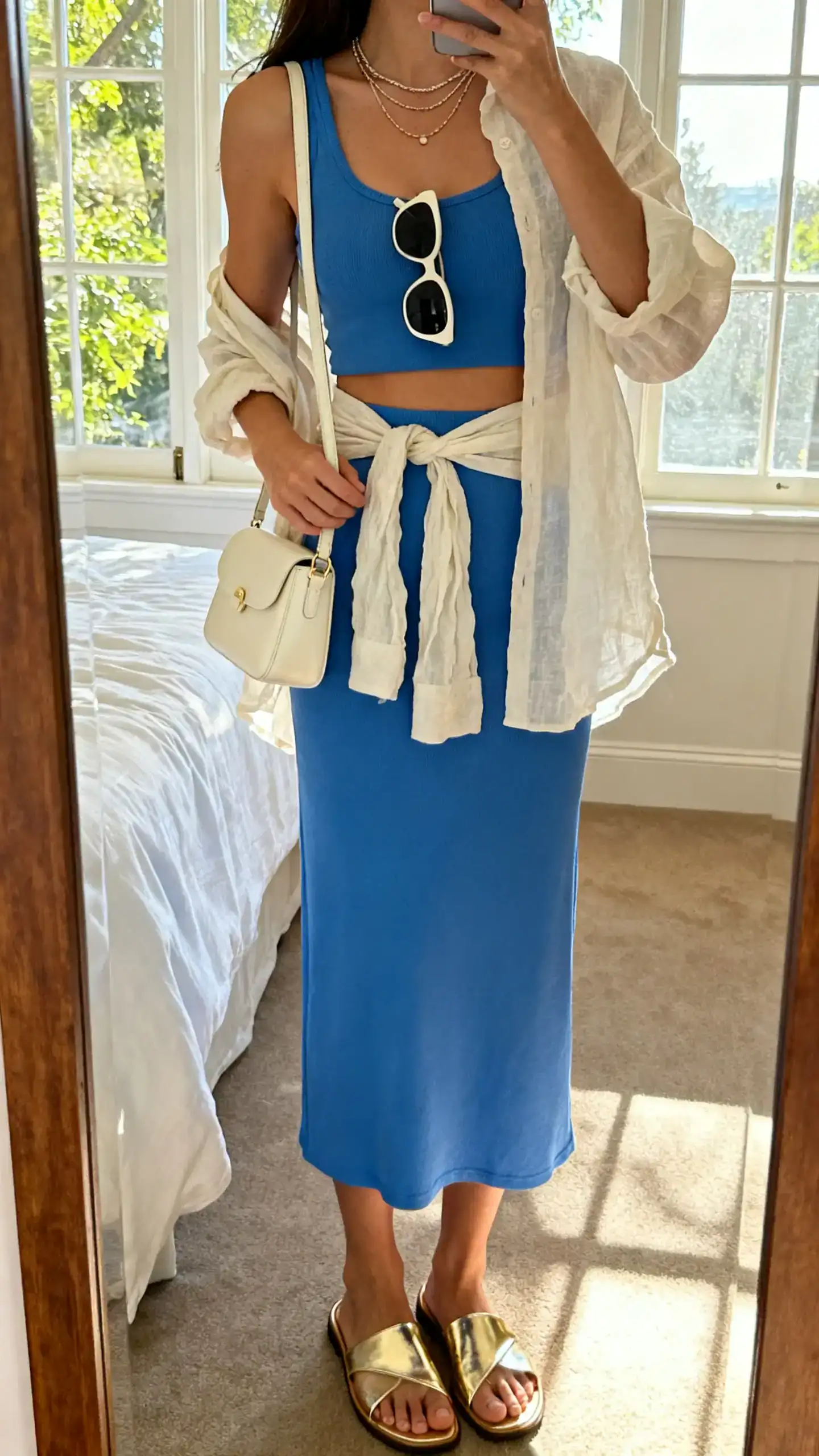 iPhone back view mirror selfie of a woman in a Capri-blue matching set (cropped tank and midi skirt), metallic gold flat sandals, delicate layered necklaces, a mini shoulder bag in cream, oversized sunglasses, and a lightweight linen shirt tied at the waist, face not visible, bedroom mirror, afternoon window light, iPhone photo quality.