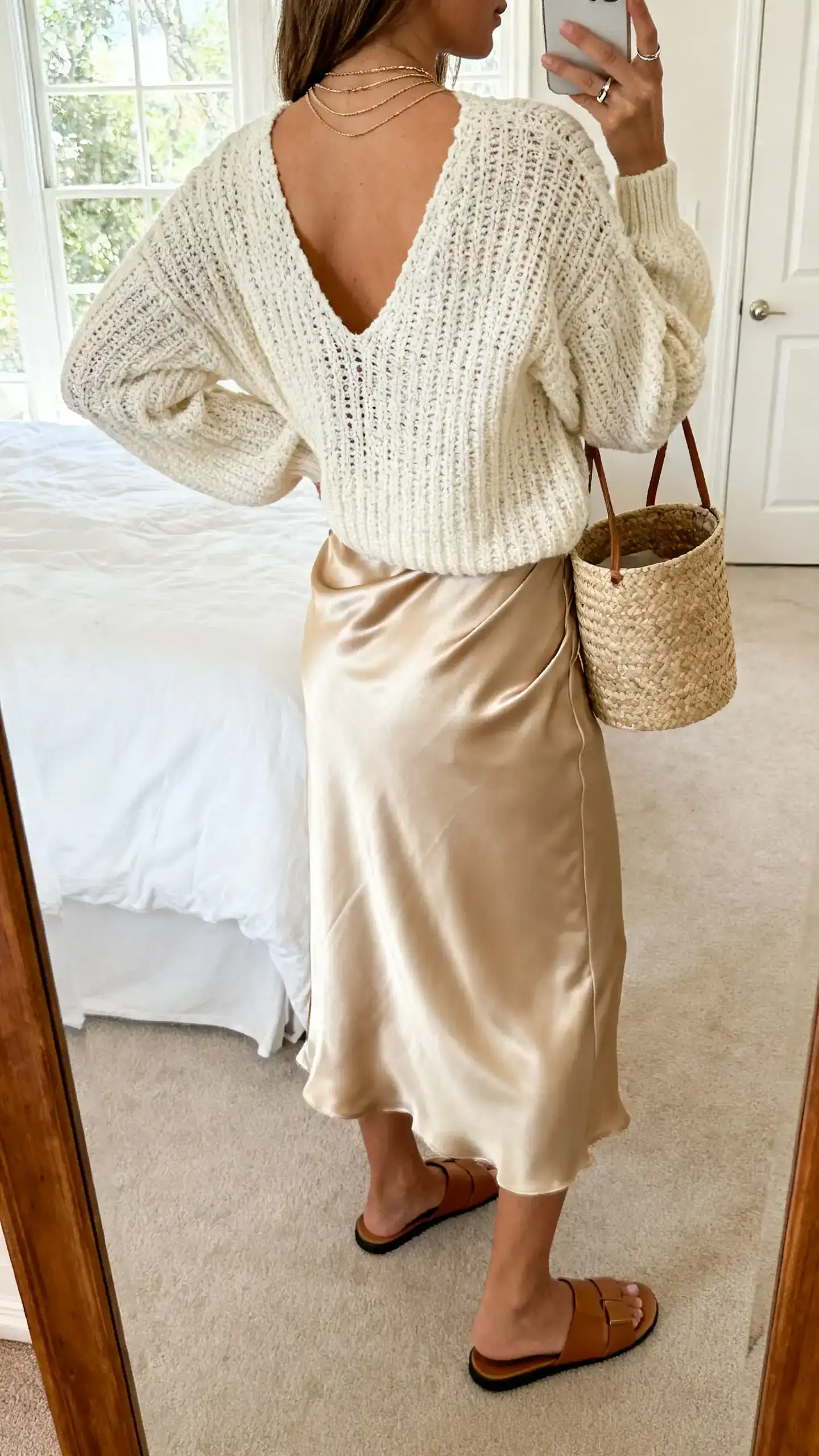 iPhone back-view mirror selfie of a woman in a cream open-knit sweater paired with a champagne satin bias-cut midi slip skirt, minimal leather slides, raffia bucket bag, thin layered necklaces and simple ring stack, face not visible, bedroom mirror, soft window light, iPhone photo quality.