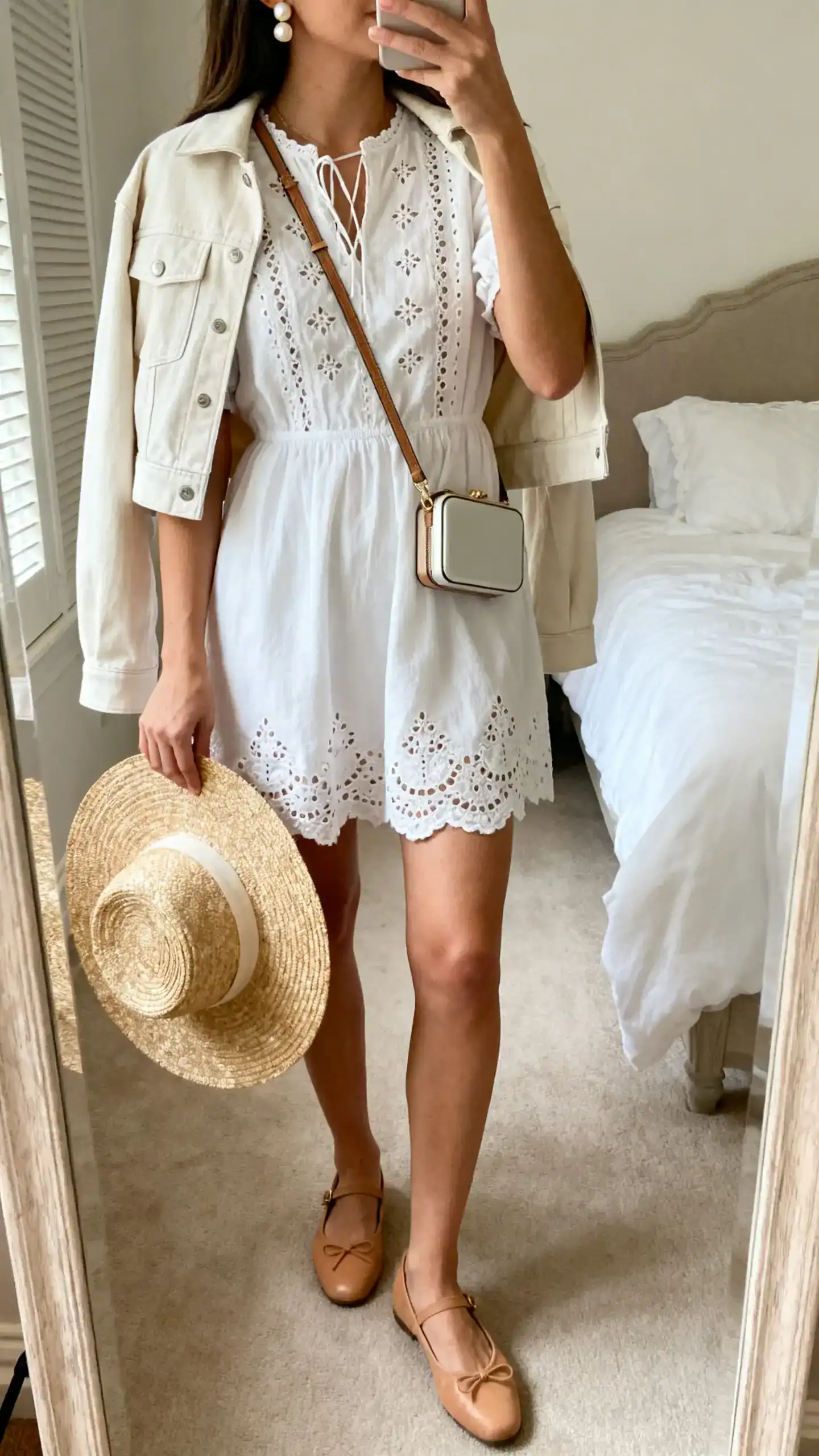 iPhone back-view mirror selfie of a woman in a white broderie anglaise mini dress, almond-toe tan ballet flats, a compact crossbody bag, pearl studs, a raffia wide-brim hat held in hand, and a light cropped jacket over shoulders, face not visible, bedroom mirror, soft morning light, iPhone photo quality.
