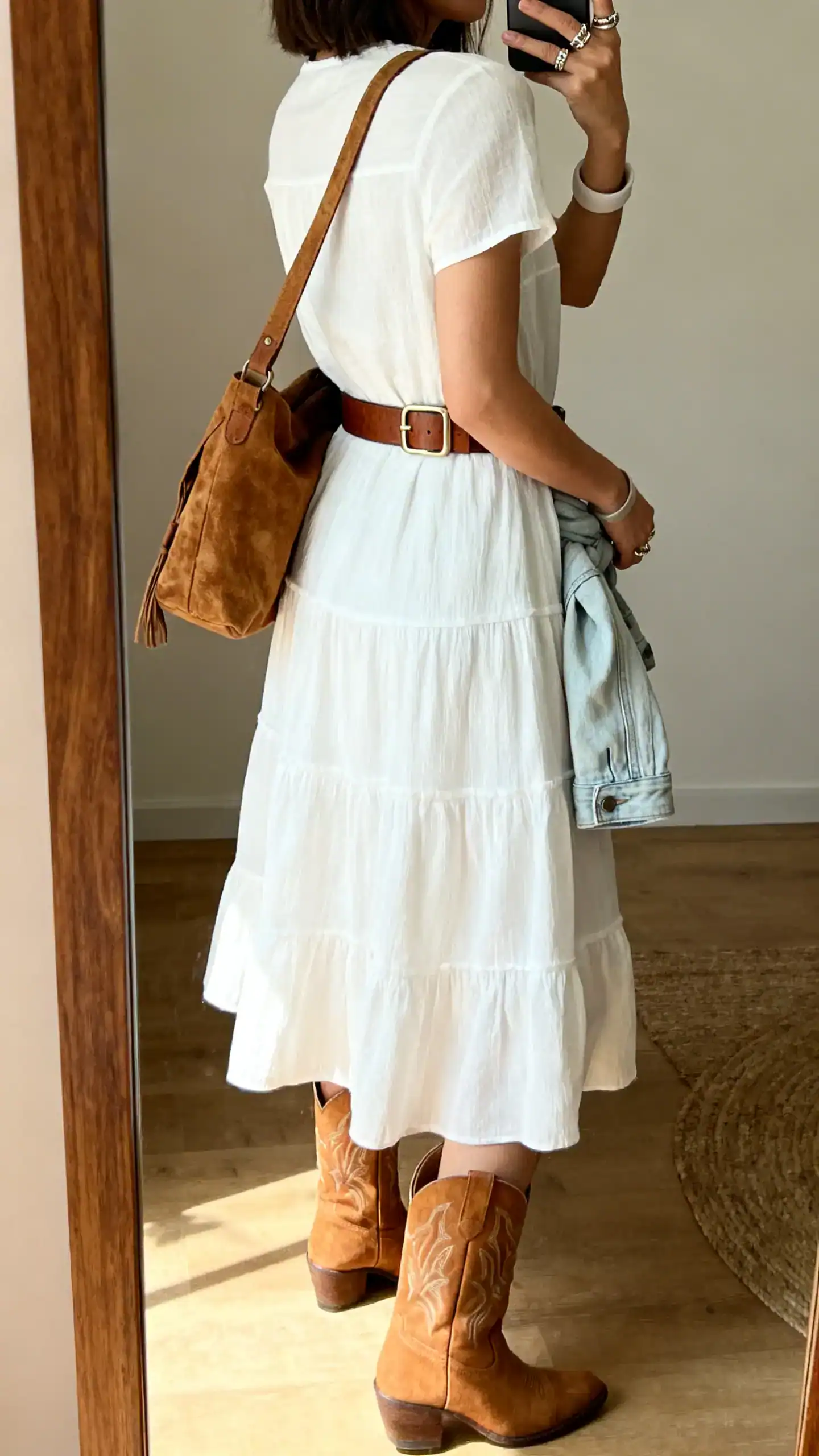 iPhone back-view mirror selfie of a woman in a white tiered midi sundress with tan western-style ankle boots, a wide leather belt with a subtle buckle, textured suede shoulder bag, stacked rings and a simple cuff, optional light denim jacket tied at waist; face not visible, full-length mirror, indoor daylight, iPhone photo quality.